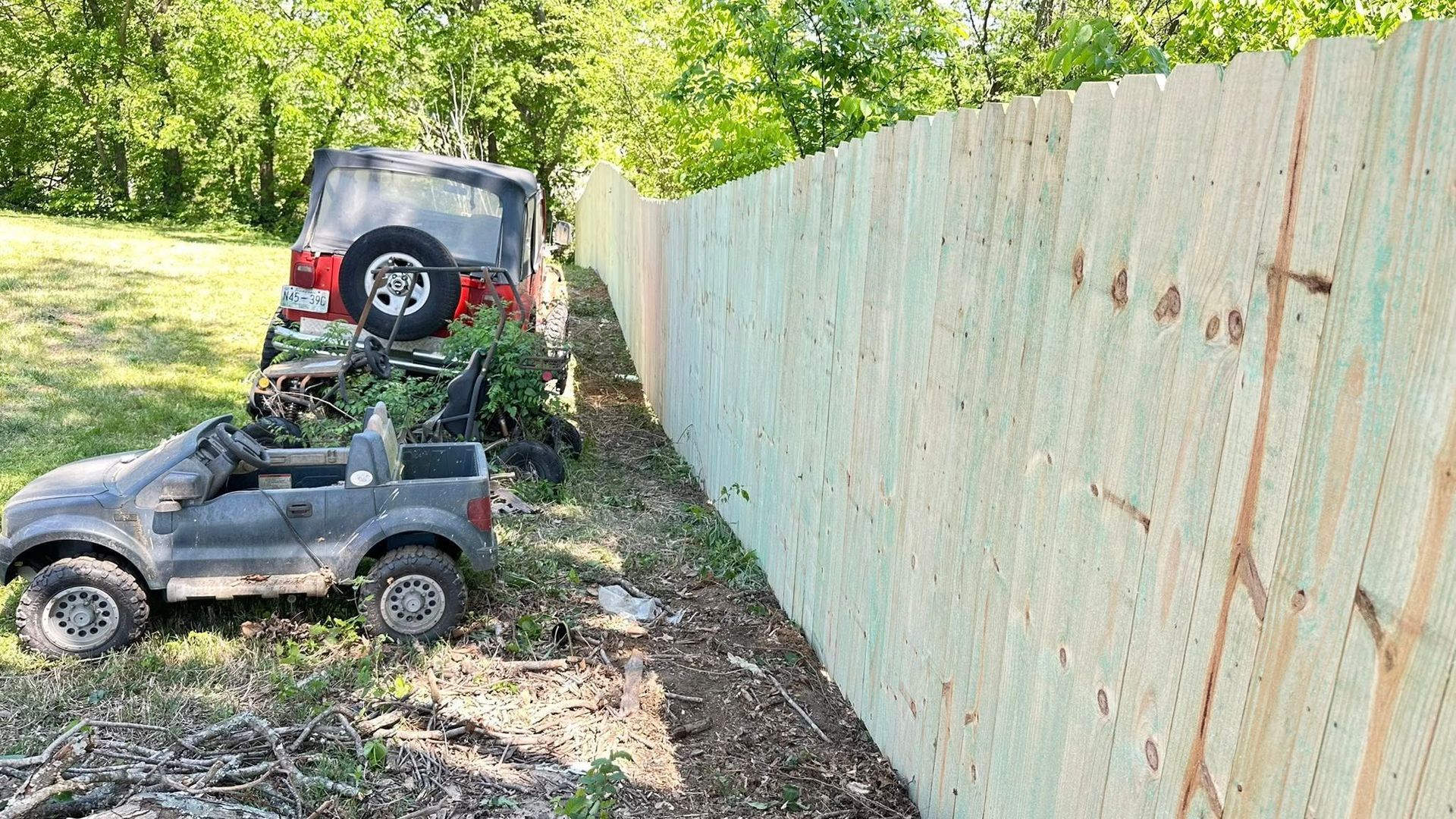 Two toy Jeeps parked beside a tall weathered fence in a grassy, wooded yard.