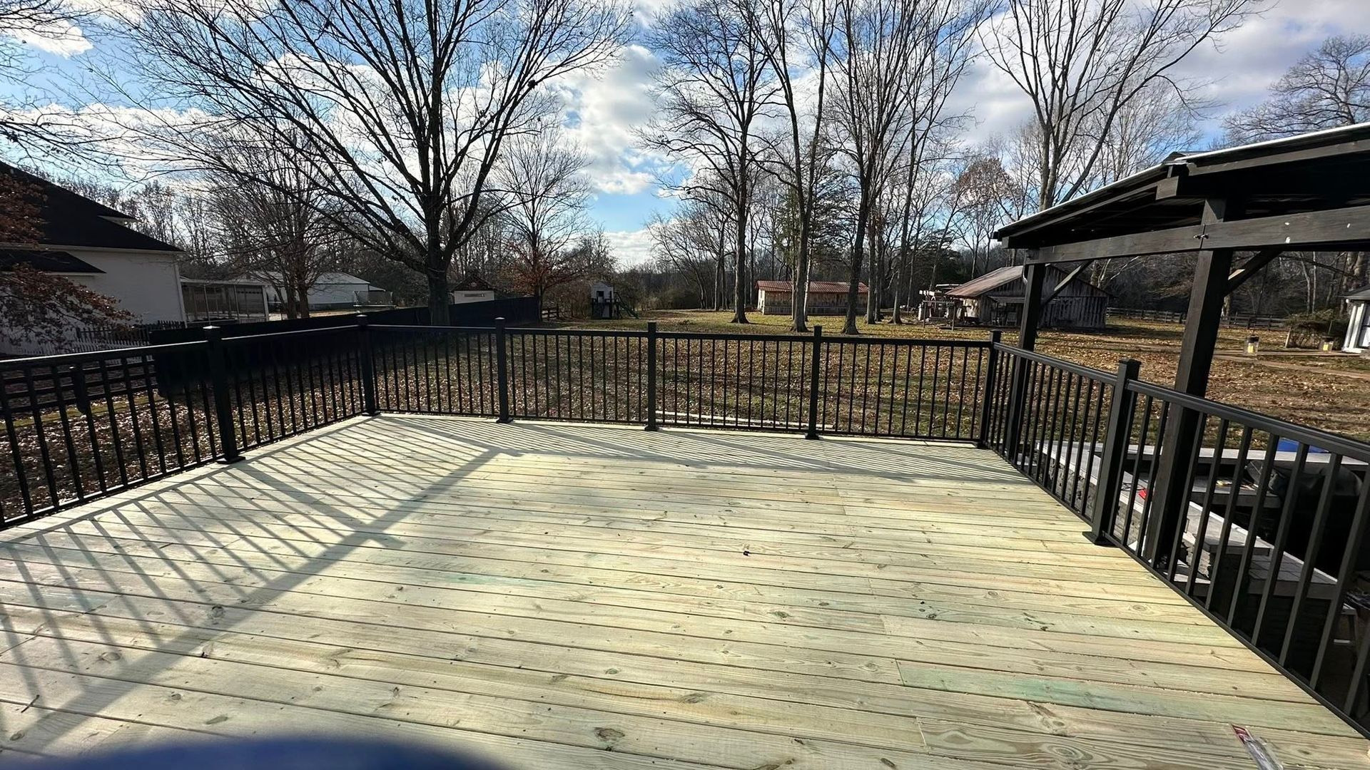 Wooden deck with black railings overlooking a yard with bare trees and neighboring homes under a bright sky