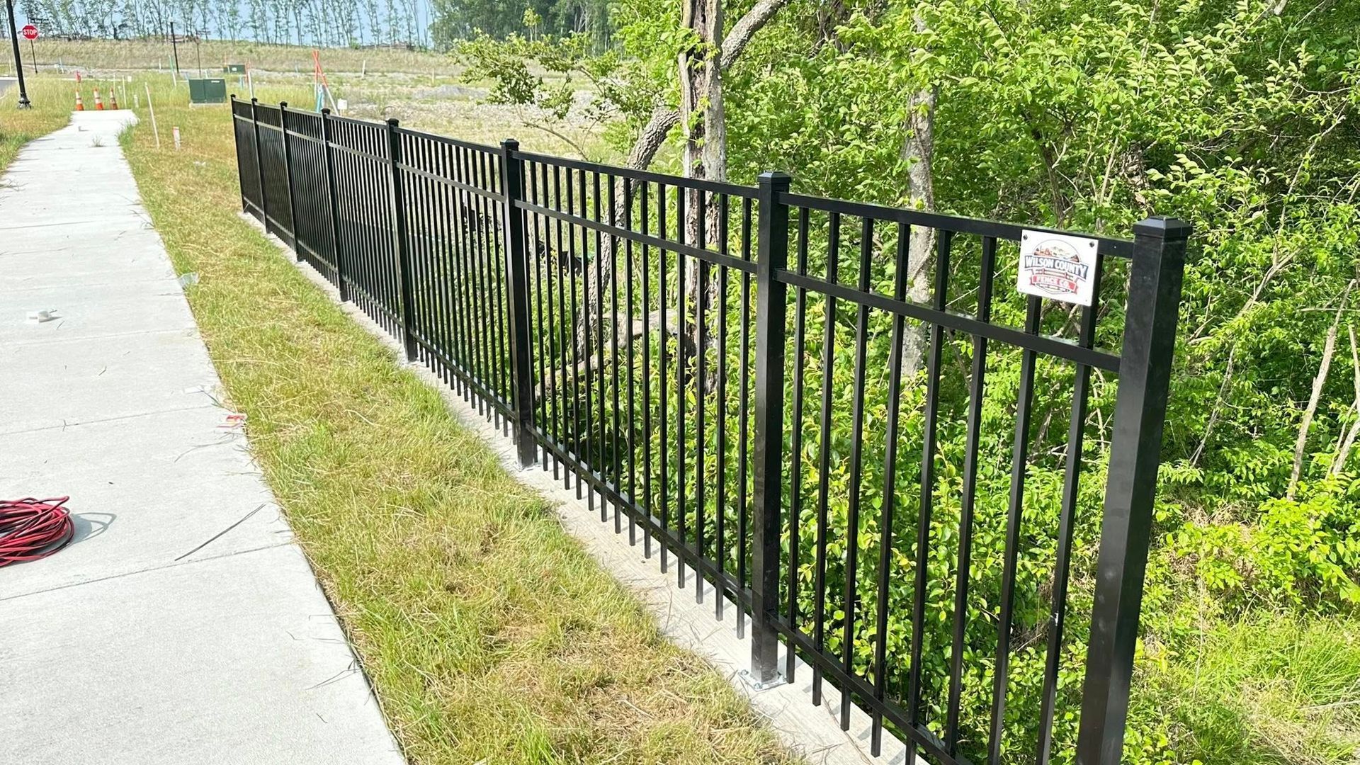 Black metal fence along a sunny sidewalk beside green bushes and grass