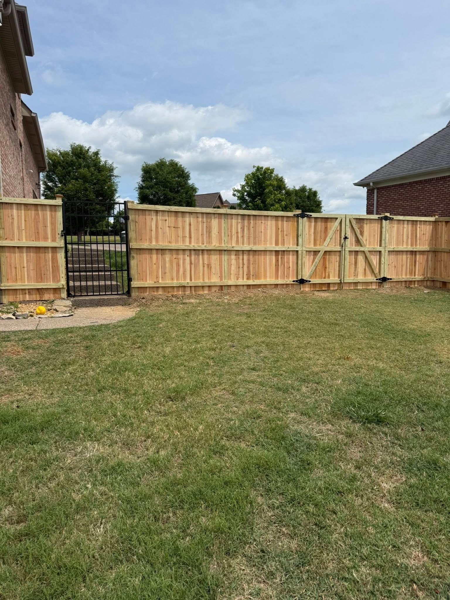 Backyard with a wooden fence, black gate, and a grassy lawn under a cloudy sky
