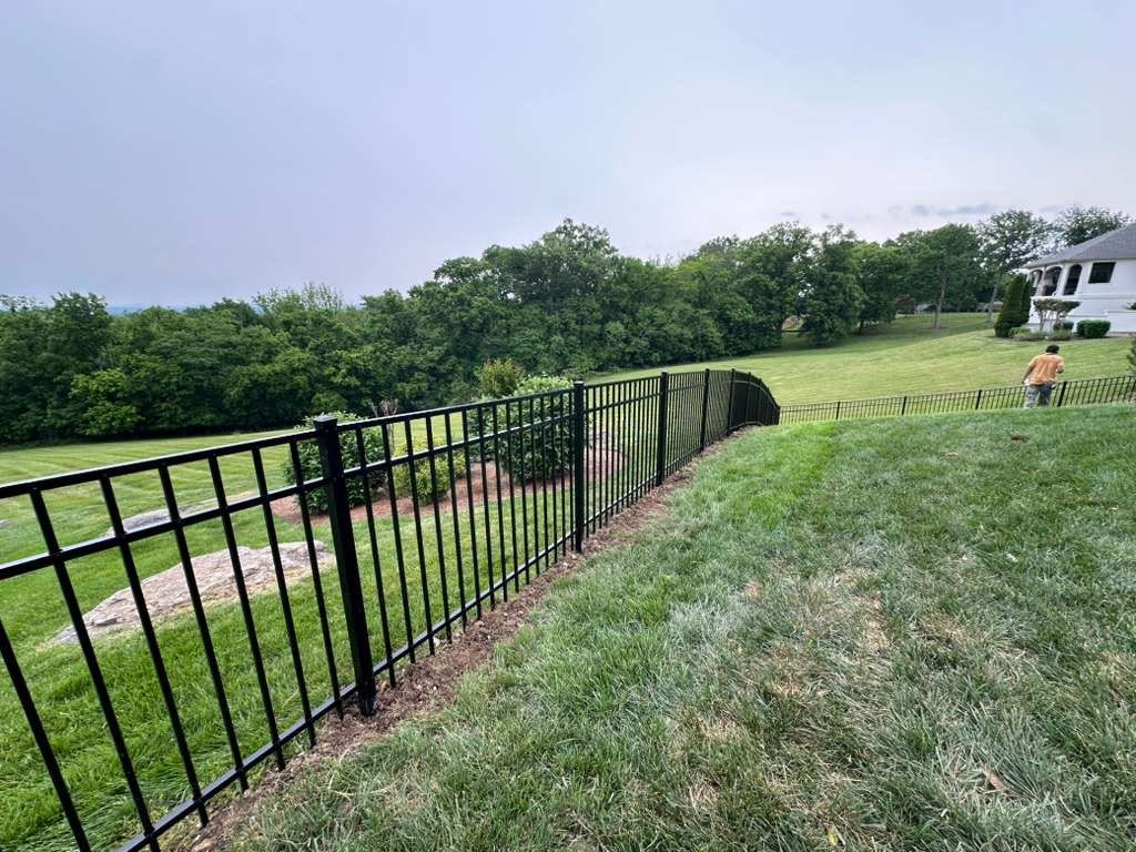 Black metal fence along a grassy slope by a wooded backyard, with a person near a house in the distance