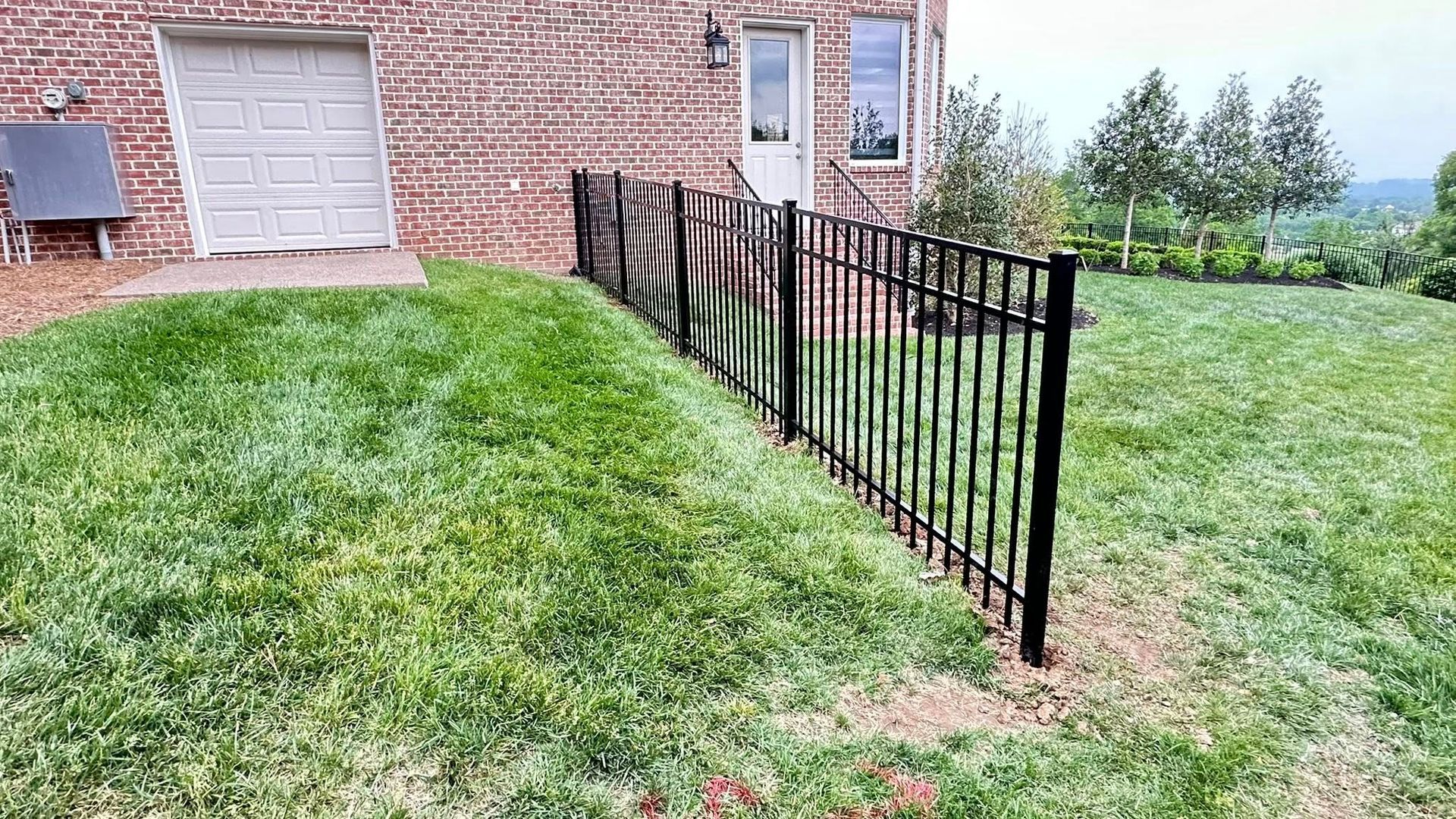 Black metal fence beside a brick house, separating a grassy yard and sloping lawn with trees beyond.