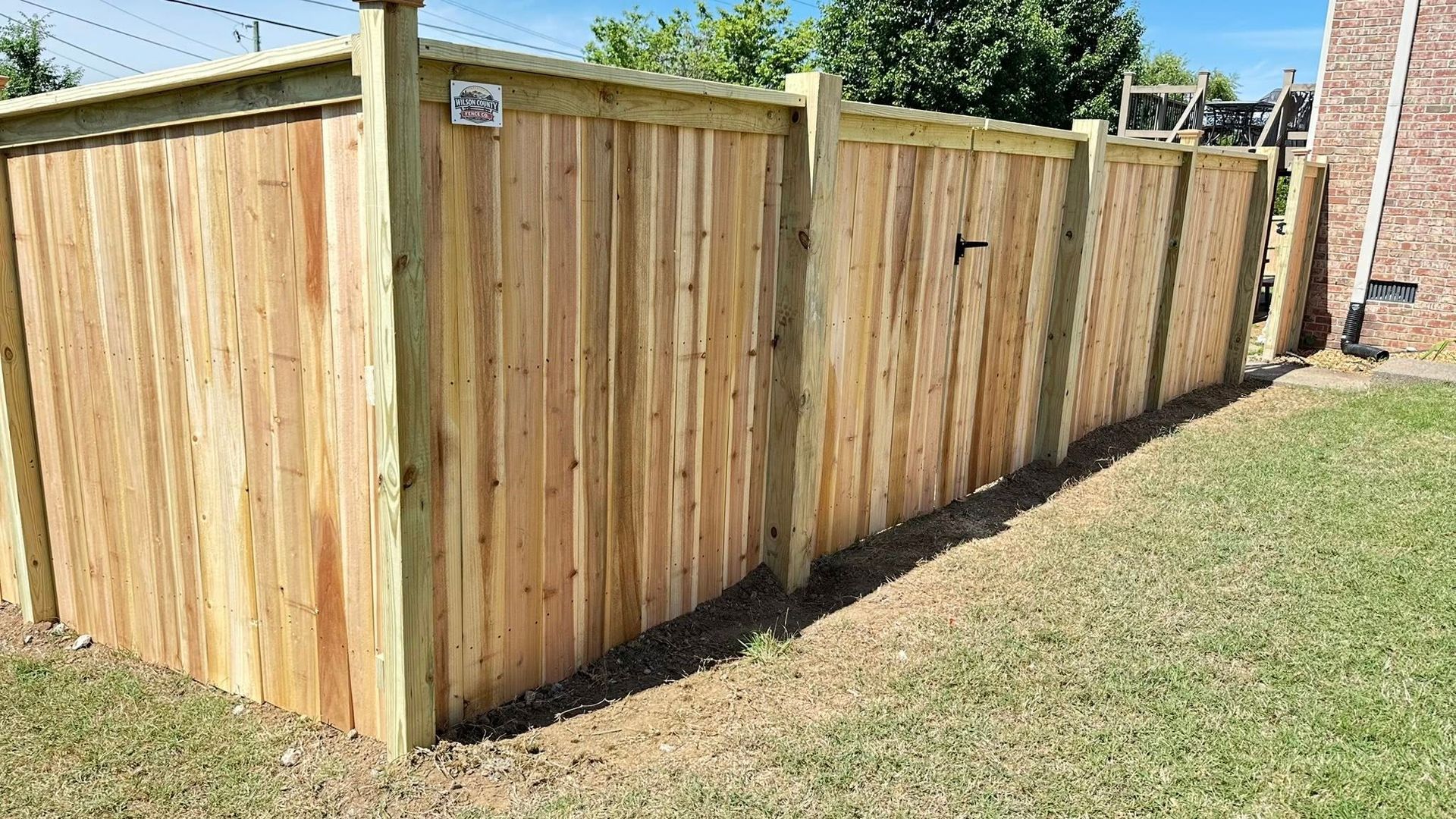 Wooden privacy fence along a grassy yard beside a brick house under a blue sky