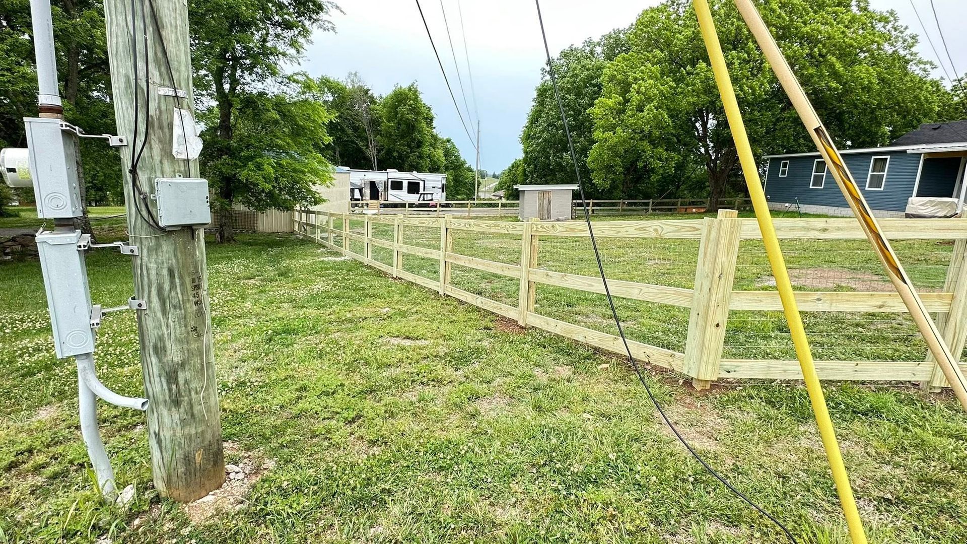 Grassy lot with a wooden fence, utility poles, and a small house in the background