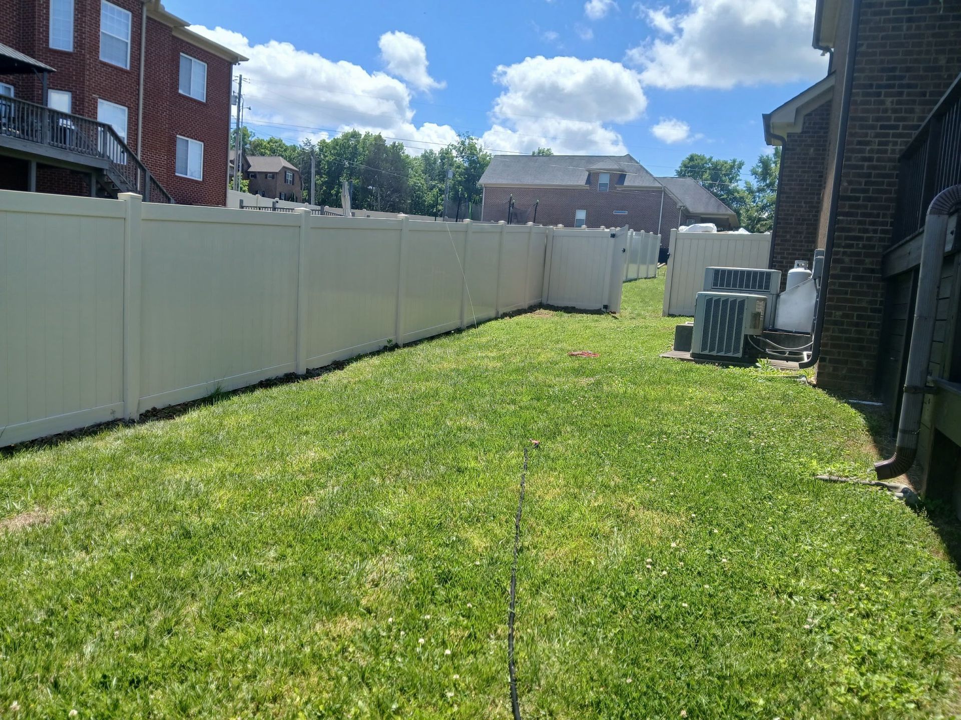 Narrow grassy backyard beside apartment buildings and a white fence under a blue sky.
