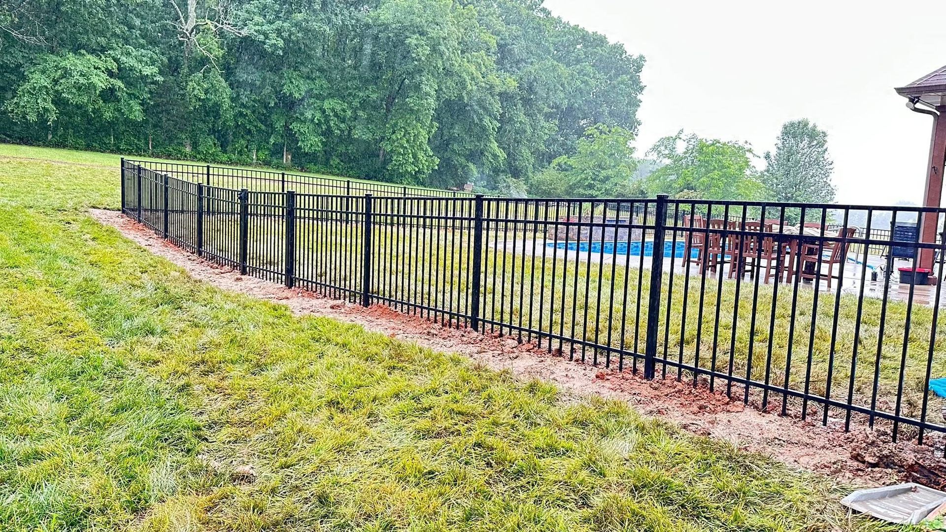 Black metal fence along a sloping grassy yard beside trees and a building on a rainy day