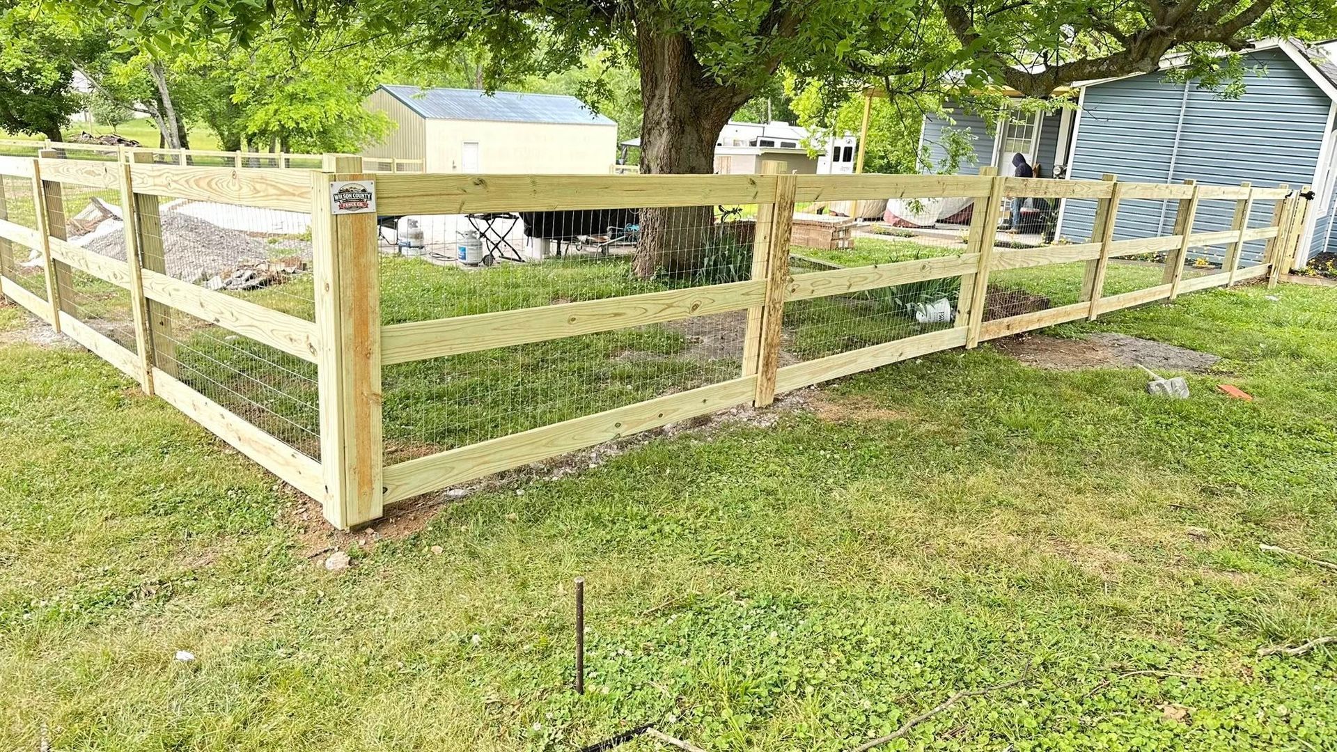 Wooden fence surrounding a grassy yard with trees, a shed, and a white vehicle in the background.