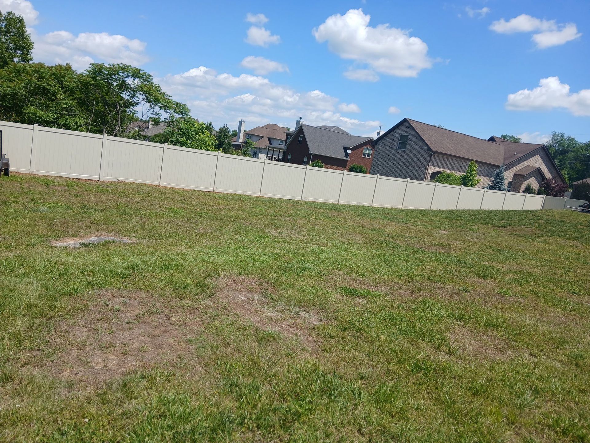Grassy vacant lot with a white fence, houses in the distance, and a blue sky with clouds.