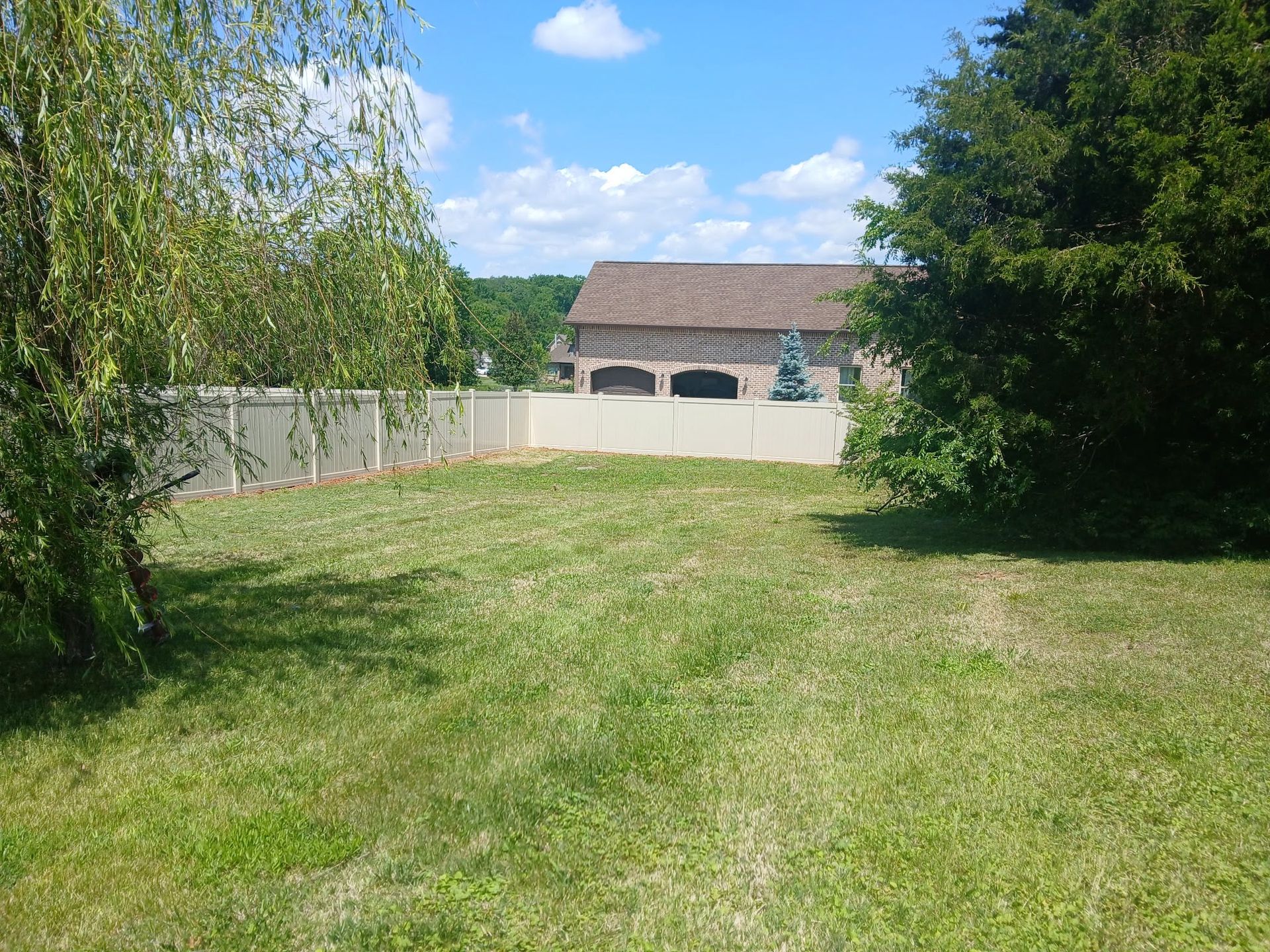 Grassy backyard with trees, a wooden fence, and a small building under a blue sky