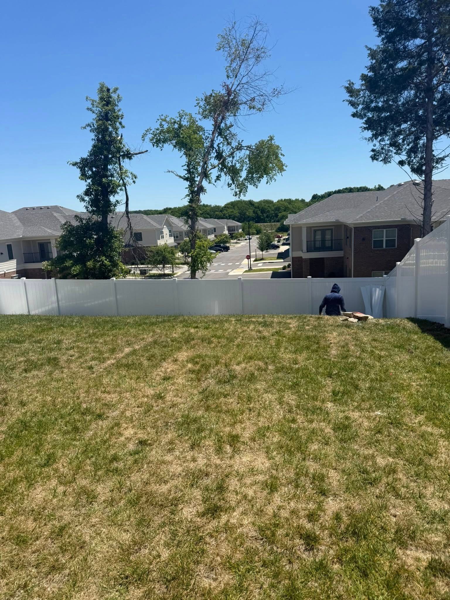 Grassy backyard with white fence, trees, and a person sitting near the fence under a blue sky