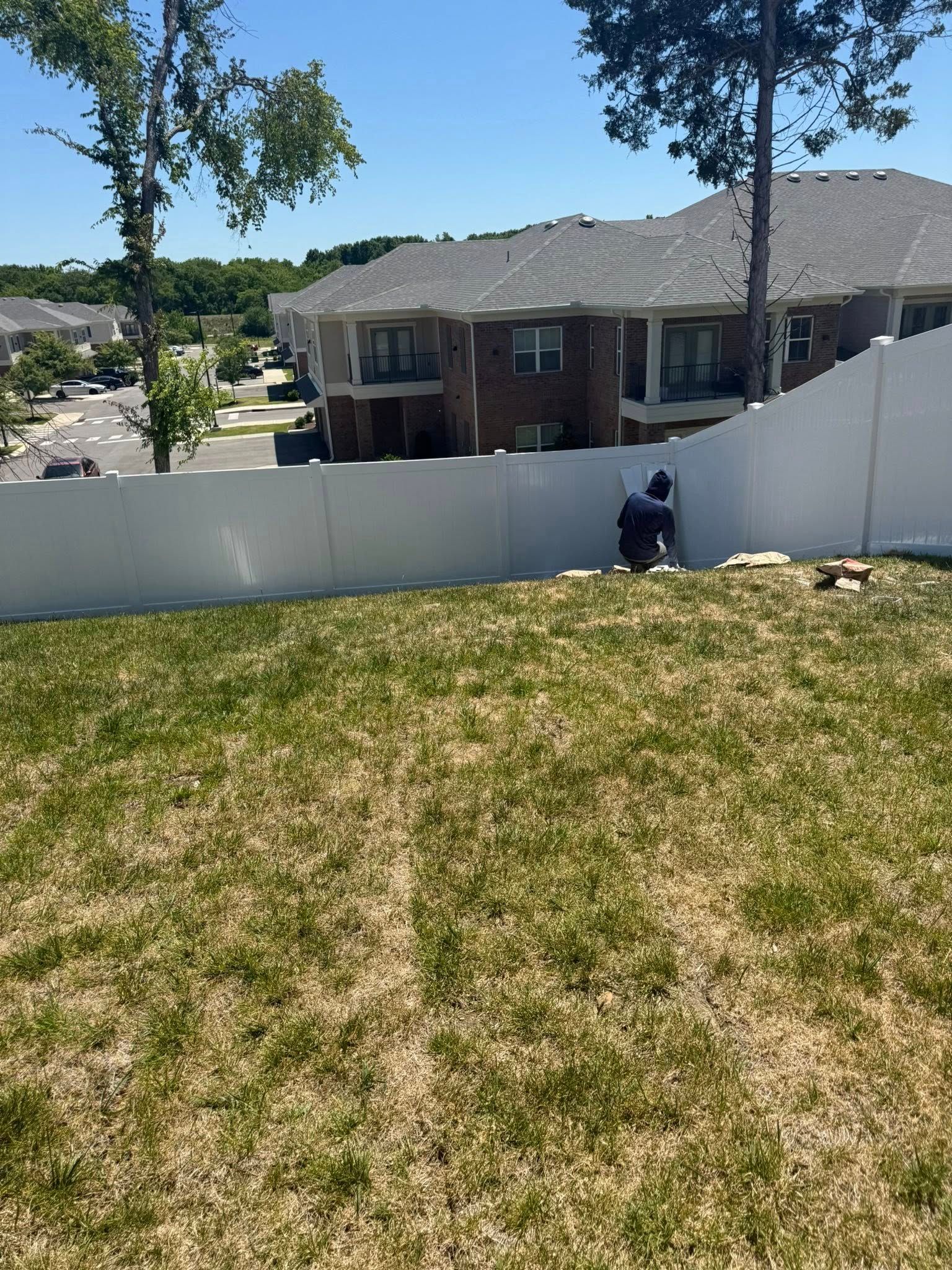 Backyard with white fence, two trees, and a brick apartment building in the background