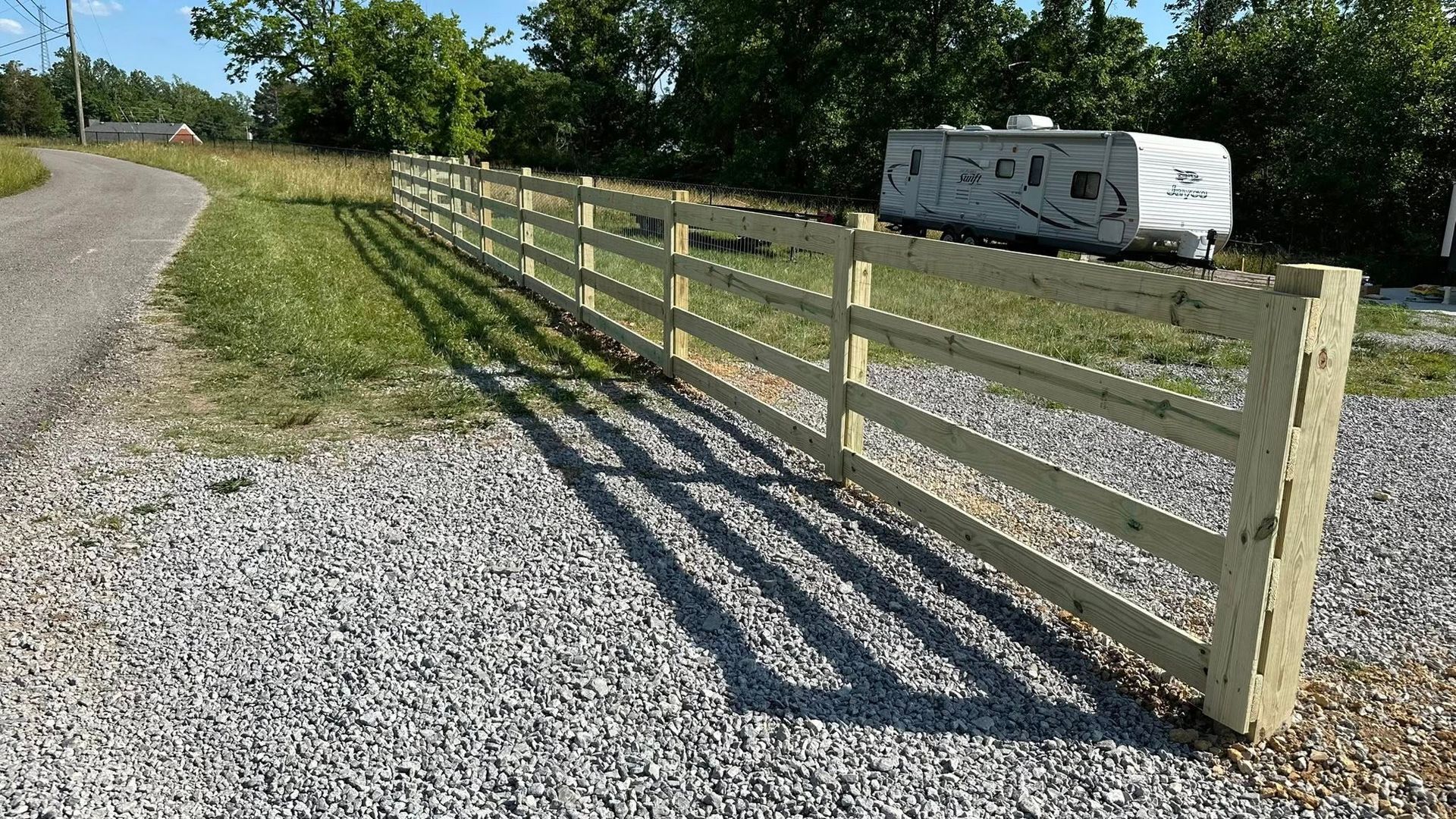 Wooden farm fence beside a gravel driveway, with a white van parked near trees in the background.