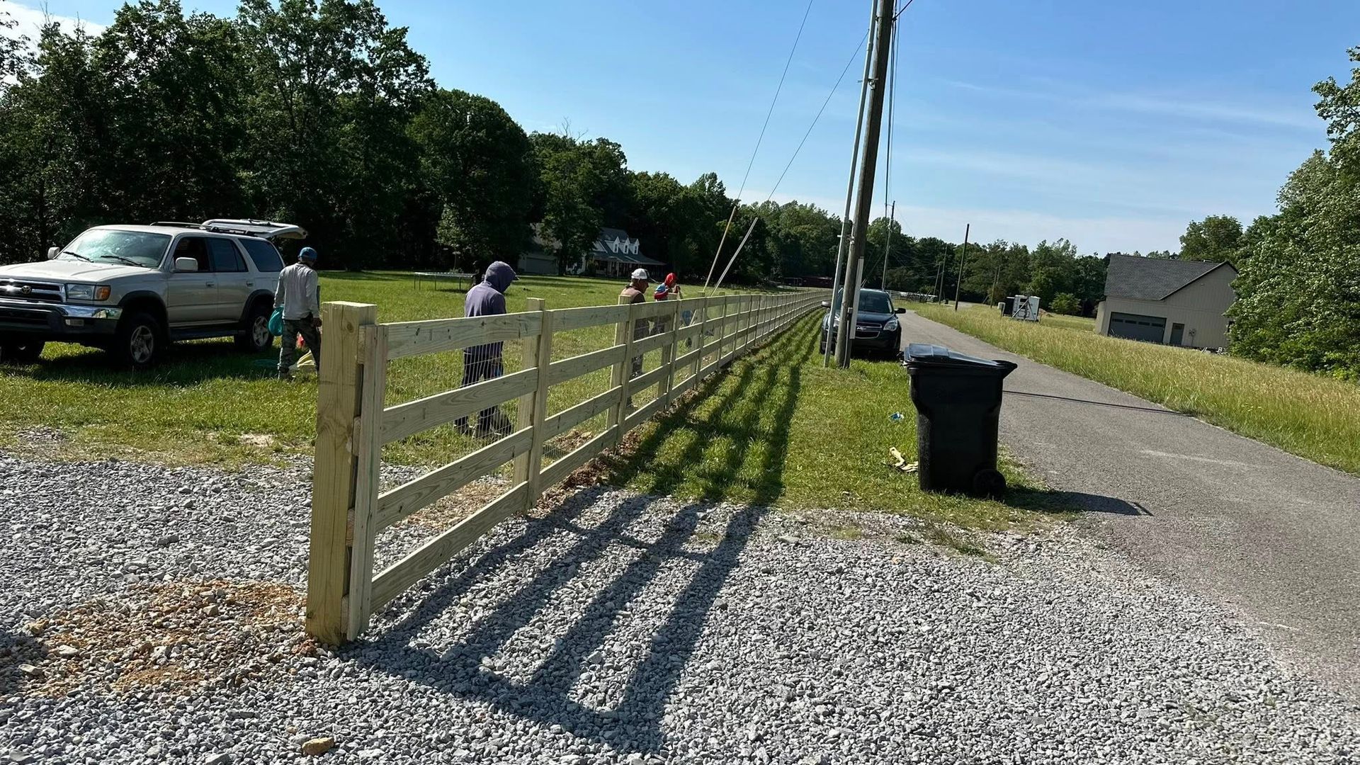 Gravel driveway with wooden fence, parked truck, trash bin, and a person in the distance on a sunny day