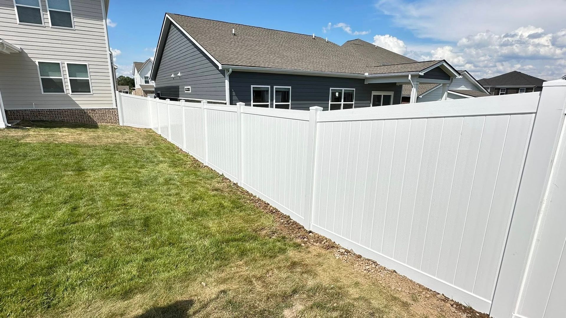 White vinyl fence bordering a grassy backyard beside gray houses under a partly cloudy sky