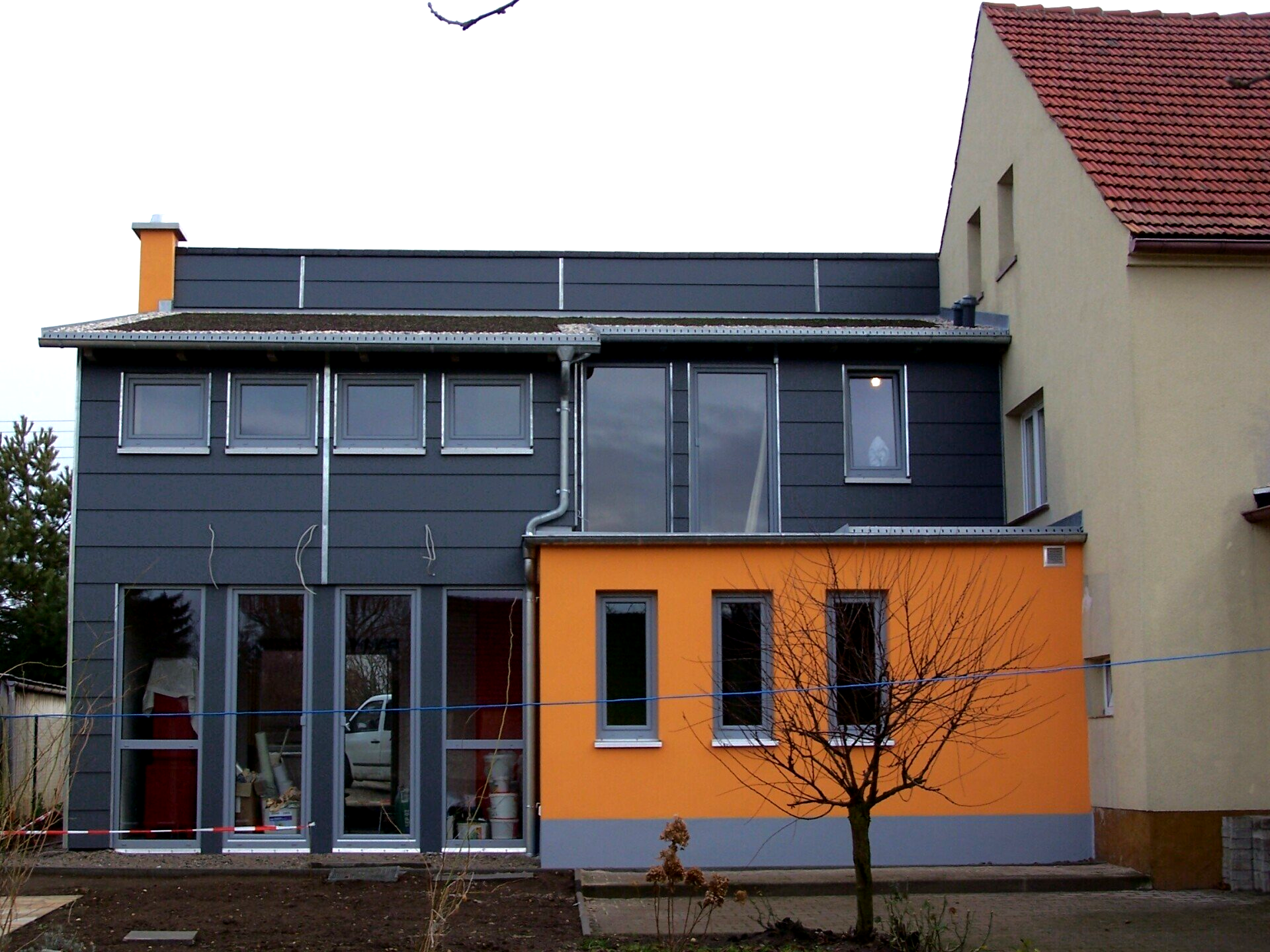 Moderner grauer und orangefarbener Gebäudeanbau mit mehreren Fenstern, neben einem hellbraunen Gebäude mit rotem Ziegeldach.