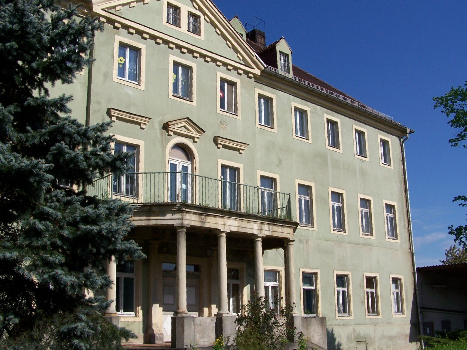 Hellgrünes Gebäude mit Balkon, Säulen und vielen Fenstern vor blauem Himmel.