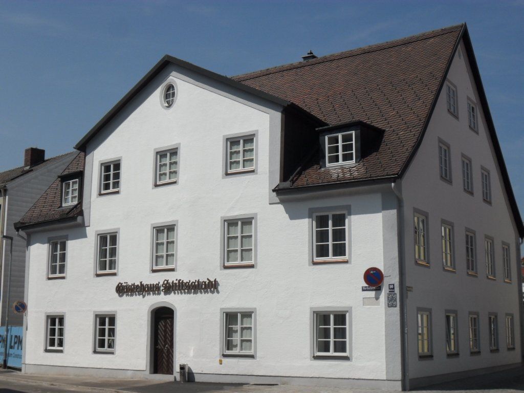 Weißes Gebäude mit braunem Dach und mehreren Fenstern. Über dem Eingang befindet sich ein Schild.