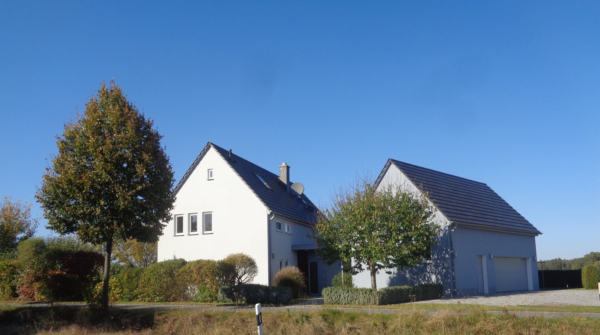 Weißes Haus mit grauem Dach und angebauter Garage, Bäume davor, klarer blauer Himmel.