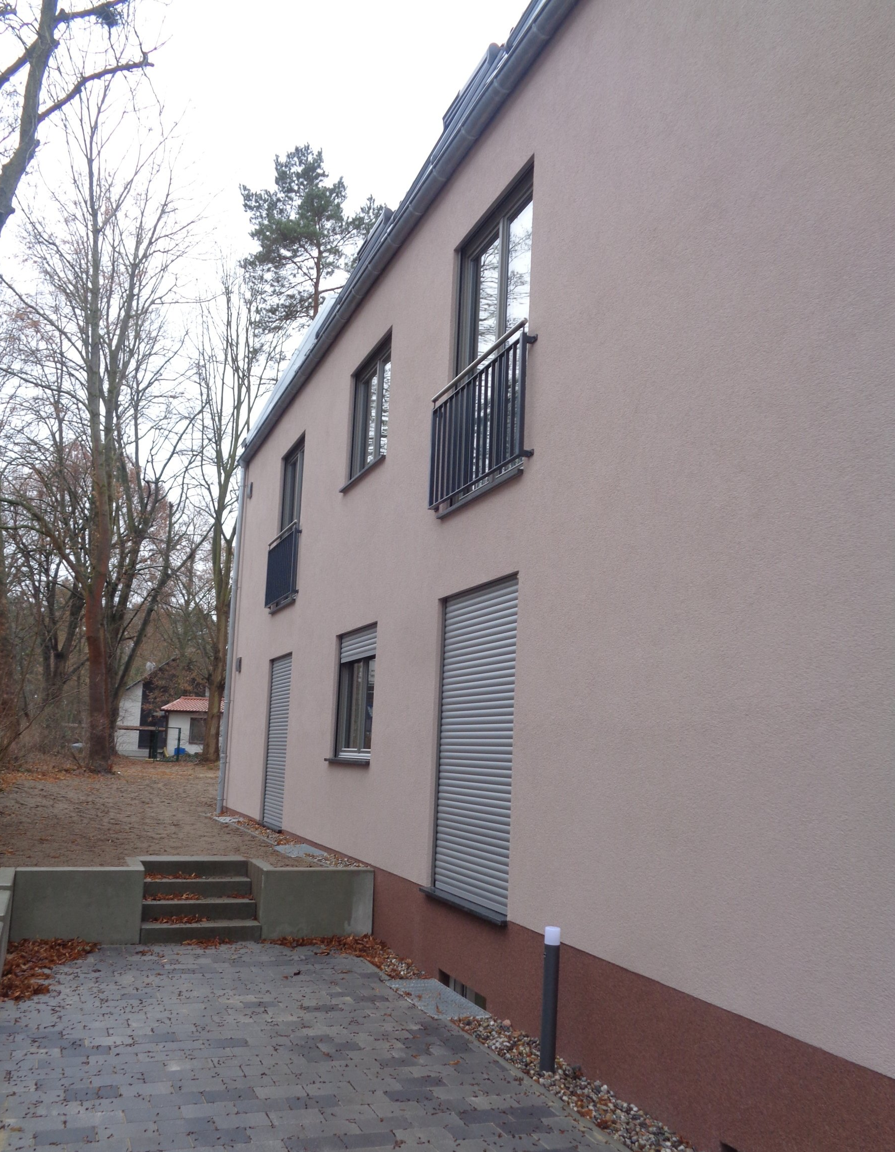 Zweistöckiges Gebäude mit beigem Stuck und dunklen Fenstern, auf einem gepflasterten Platz mit Treppe.