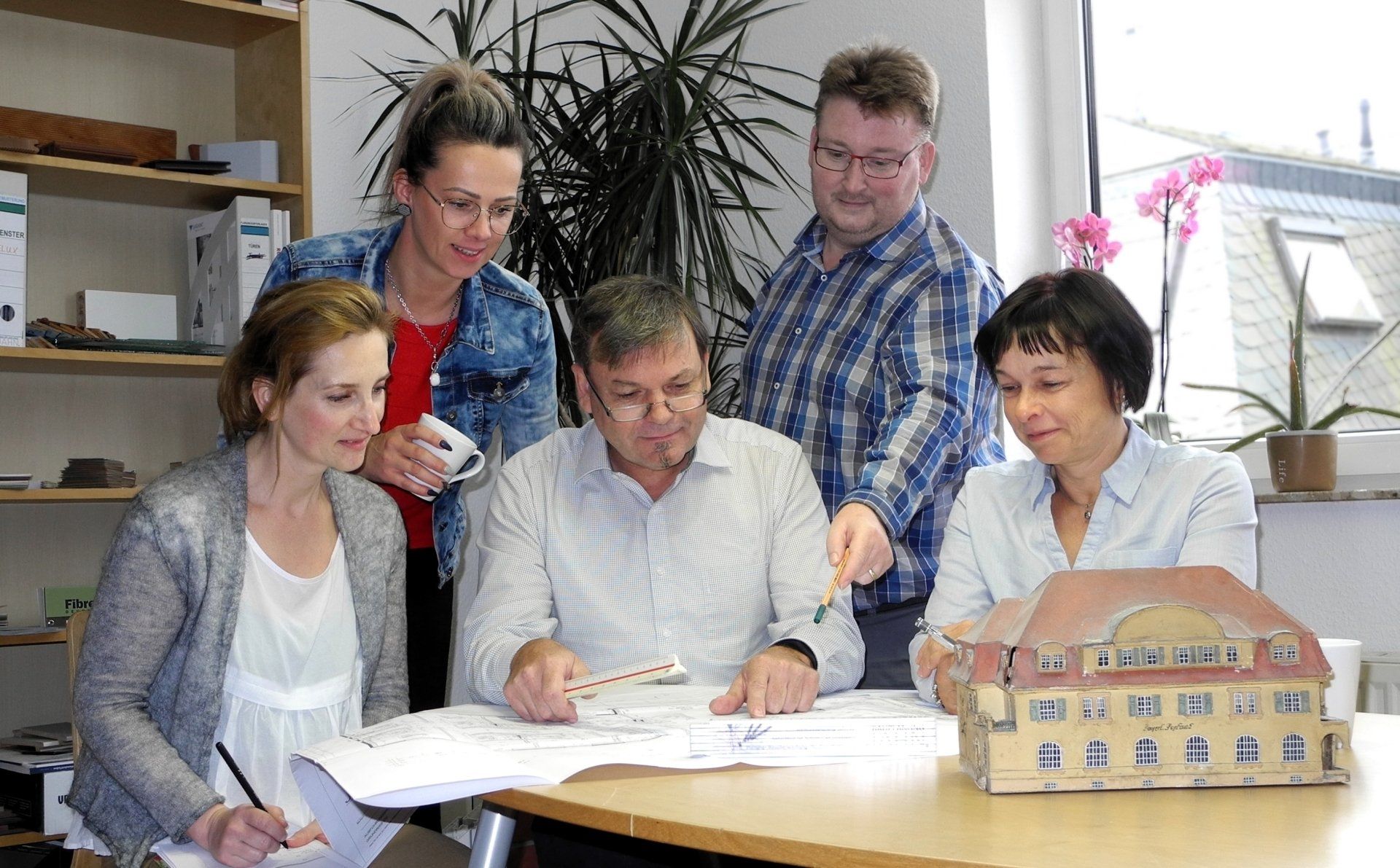 Fünf Personen sitzen an einem Tisch und prüfen Baupläne; in einem Büro befinden sich ein Modellgebäude und Dokumente.