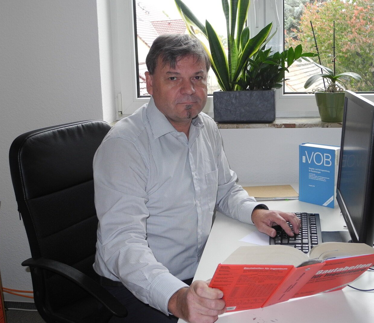 Ein Mann sitzt am Schreibtisch und tippt auf der Tastatur. Vor ihm stehen Buch und Monitor. Hinter ihm steht eine Pflanze im Fenster.