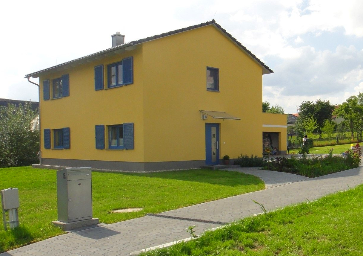 Gelbes zweistöckiges Haus mit blauen Fensterläden und Tür, auf einem grünen Rasen mit gepflastertem Gehweg.