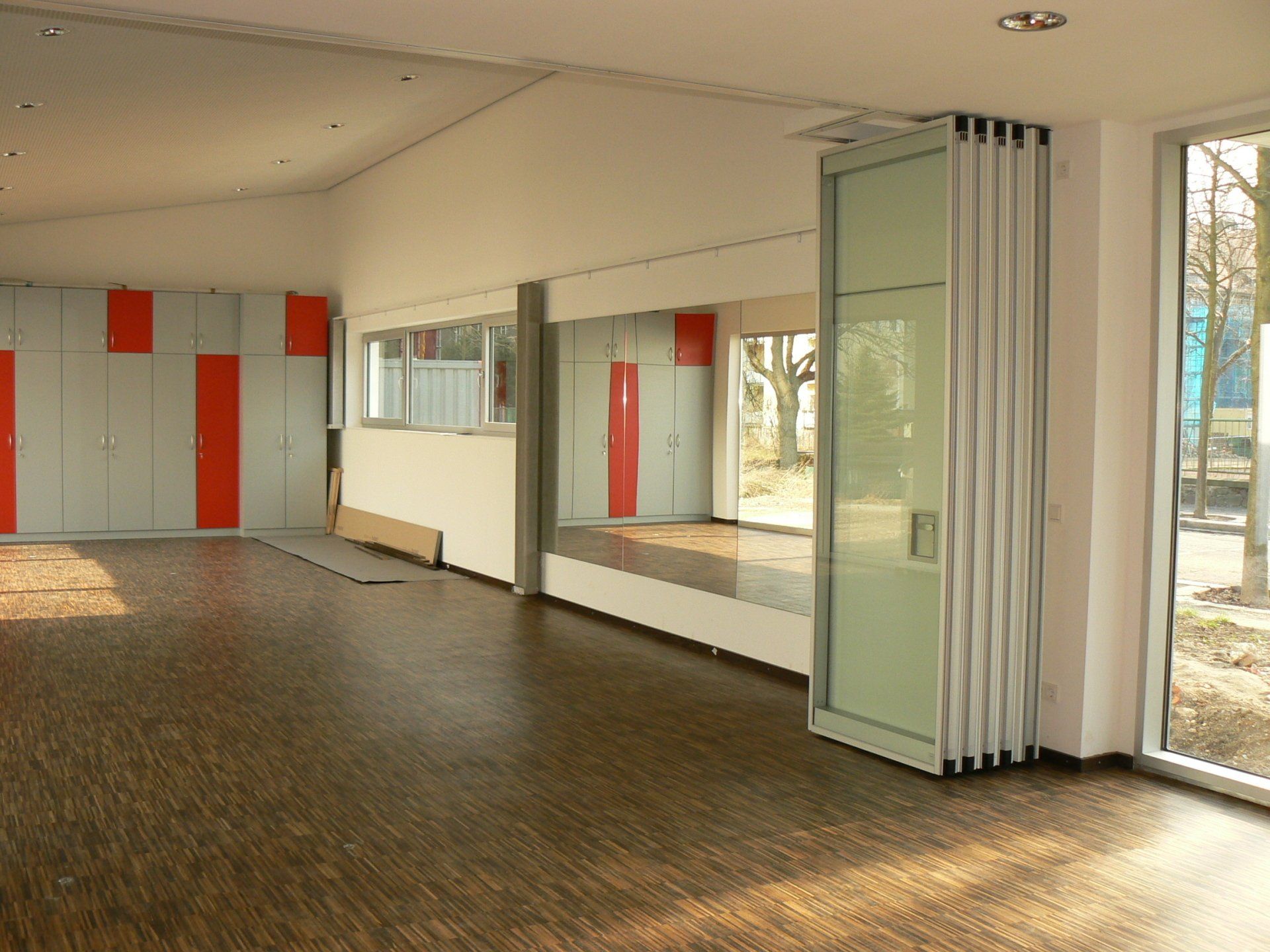 Leerer Raum mit dunklem Holzboden, weißen Wänden und einer Falttür aus Glas, mit roten und grauen Akzenten.