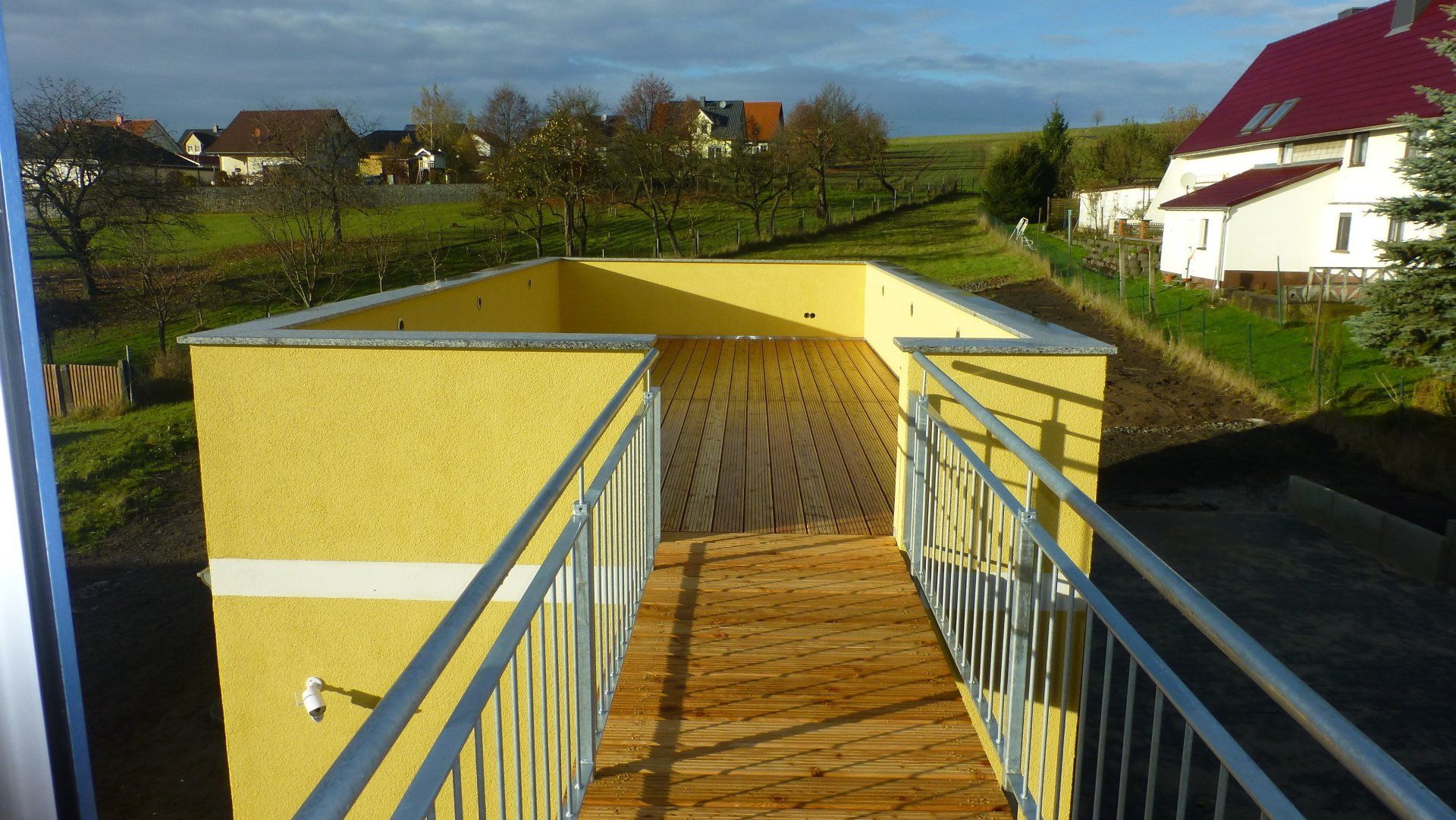 Ein gelb ummauerter, erhöhter Gehweg mit Holzboden und Metallgeländer verbindet zwei Bereiche.