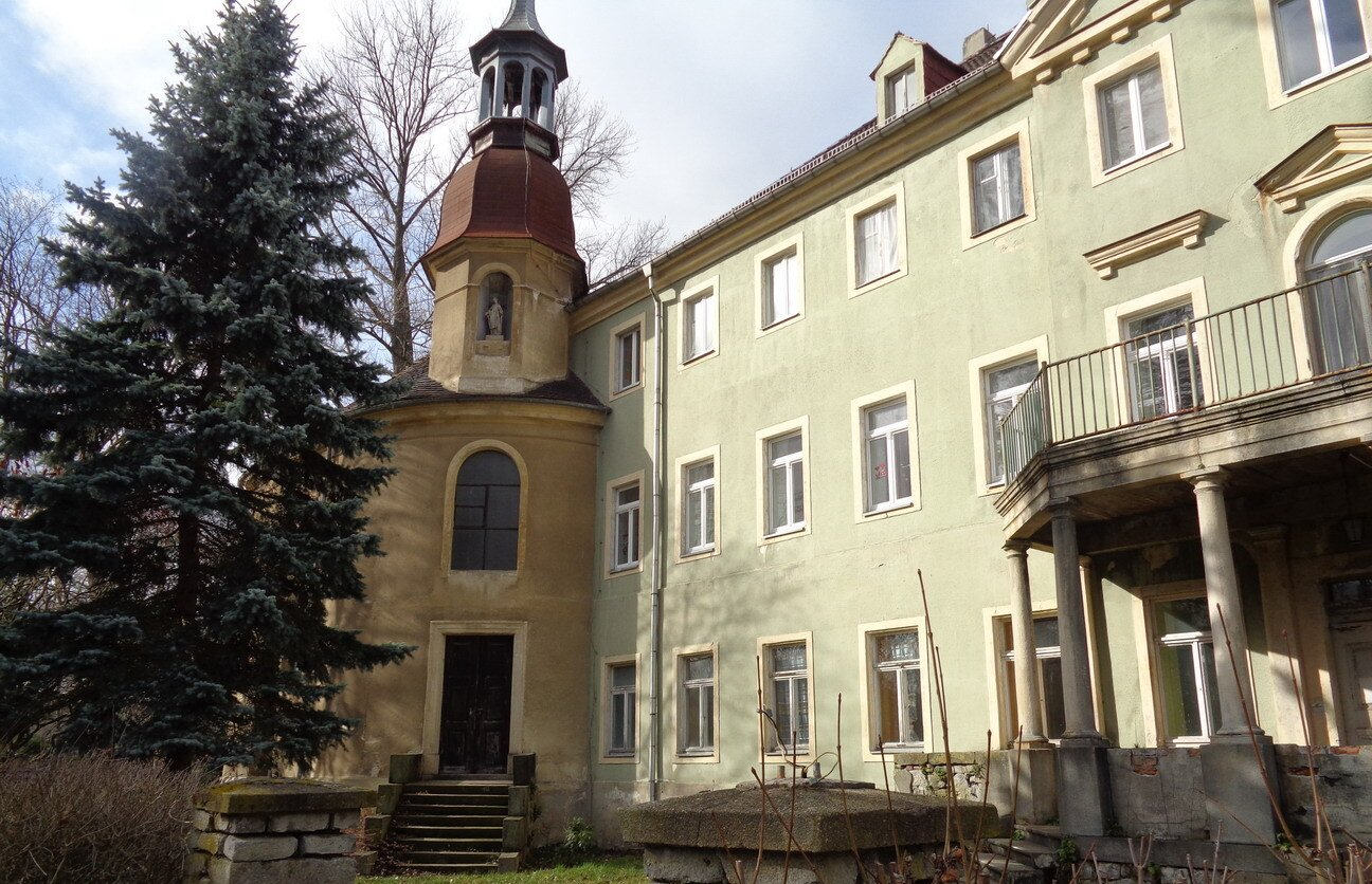 Ein hellgrünes Gebäude mit einem Turm. Links steht ein hoher immergrüner Baum.