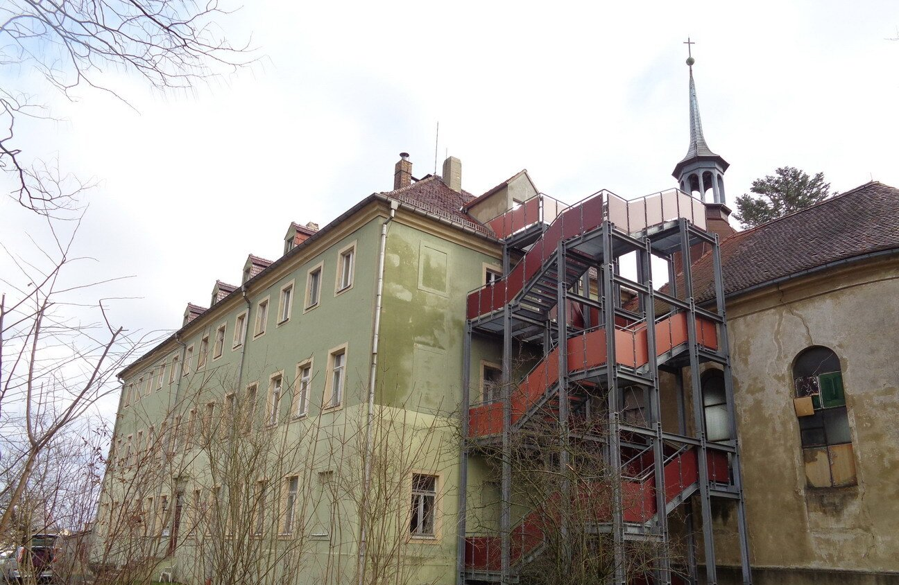Ein verwittertes Gebäude mit einer außenliegenden Metalltreppe und einem Kirchturm vor einem bewölkten Himmel.