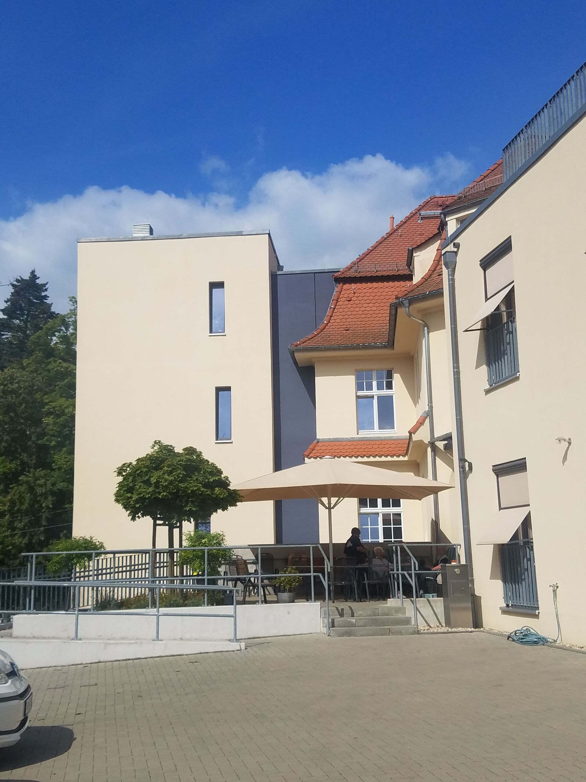 Außenansicht eines beigefarbenen Gebäudes mit rotem Ziegeldach, Terrasse und blauem Himmel.