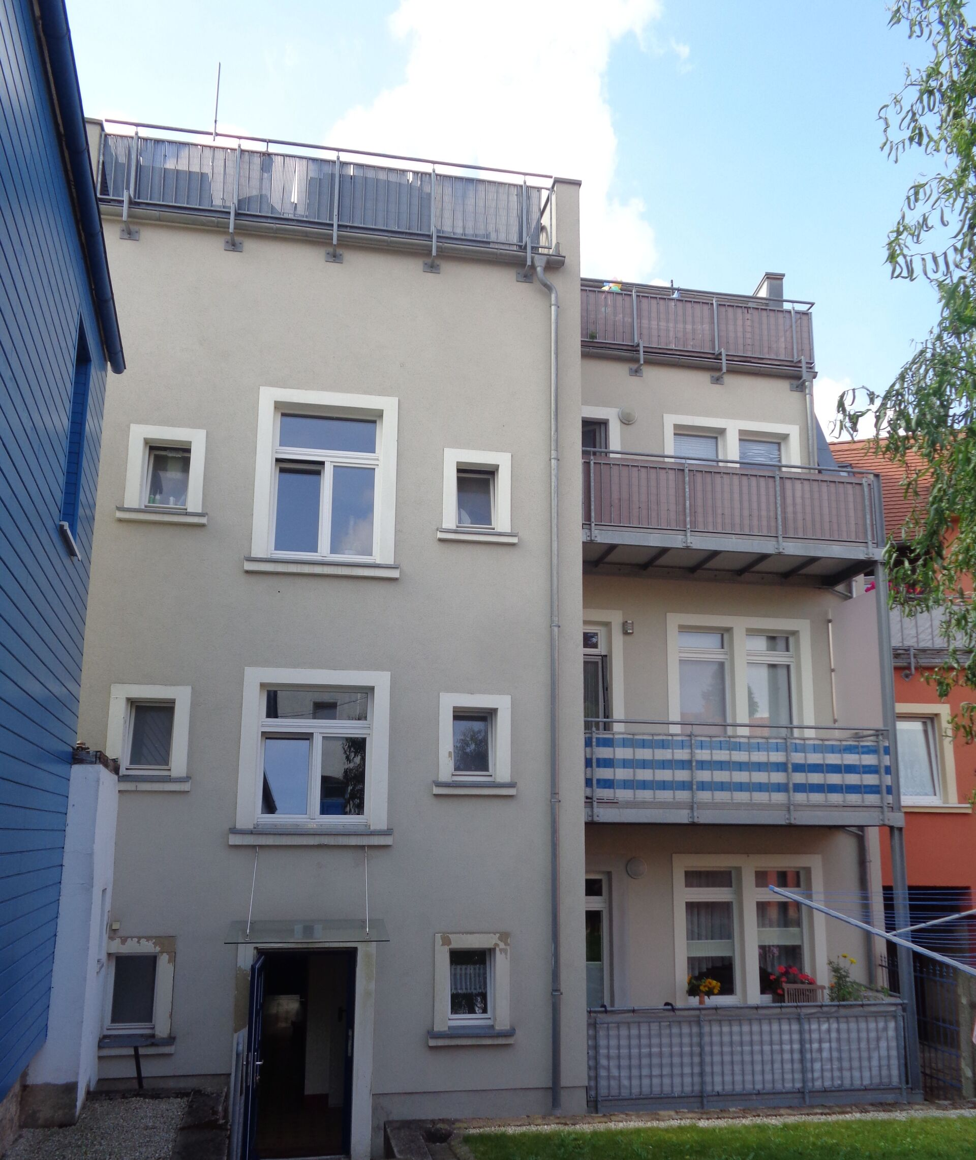 Beigefarbenes Wohnhaus mit Balkonen, kleinen Fenstern und dunklem Eingang, von der Seite gesehen.