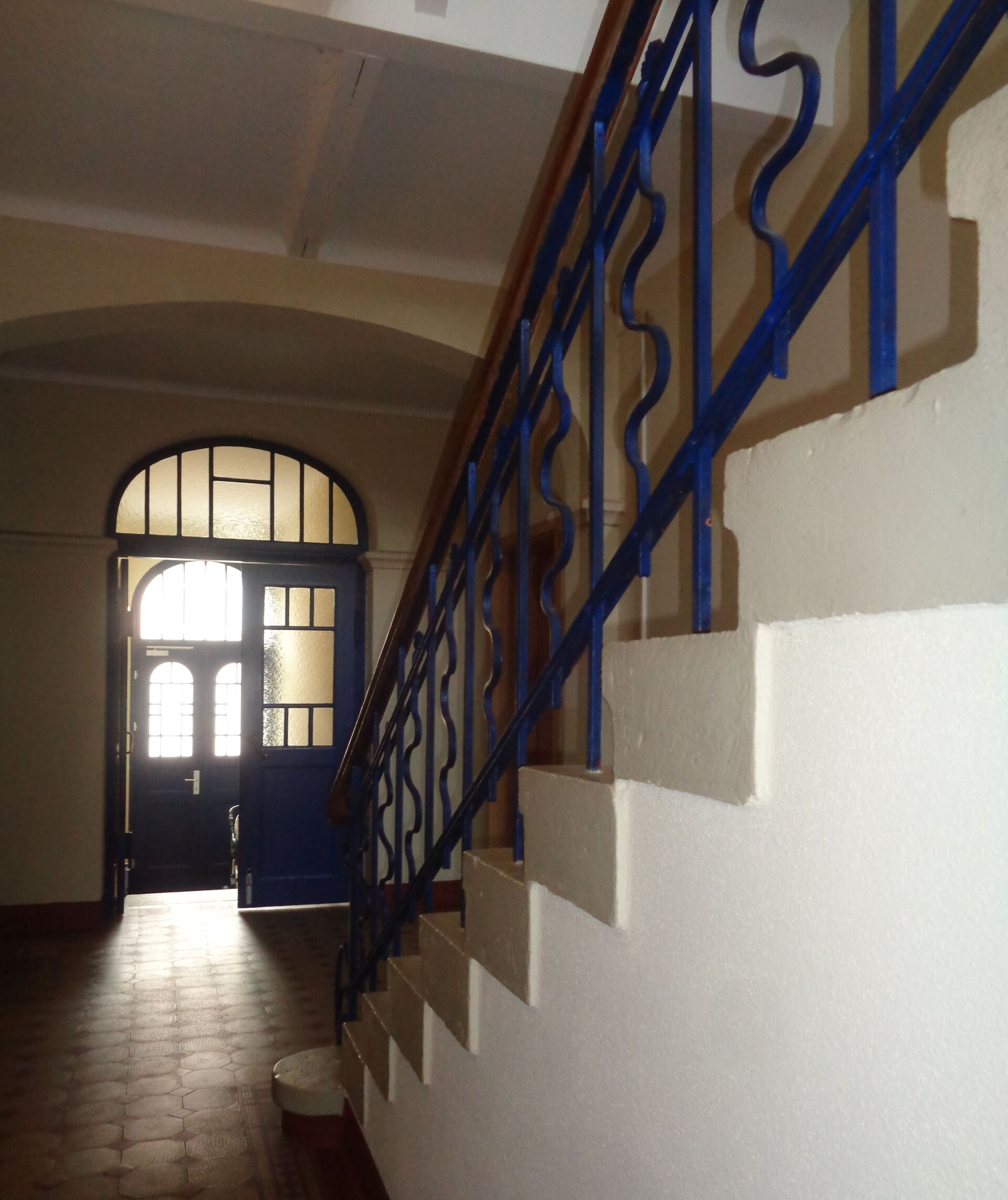 Innenflur mit Treppe, blauem Geländer und gewölbter Tür, durch die Sonnenlicht fällt.