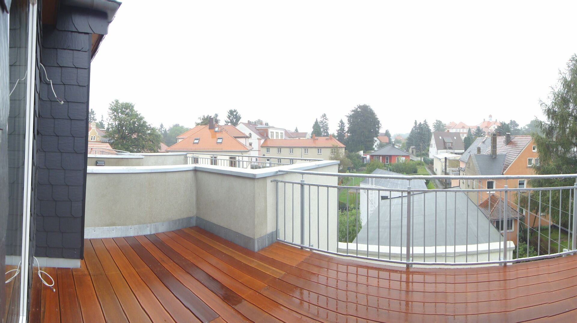 Dachterrasse mit Holzboden, Metallgeländer und Blick auf Gebäude und Bäume unter bewölktem Himmel.