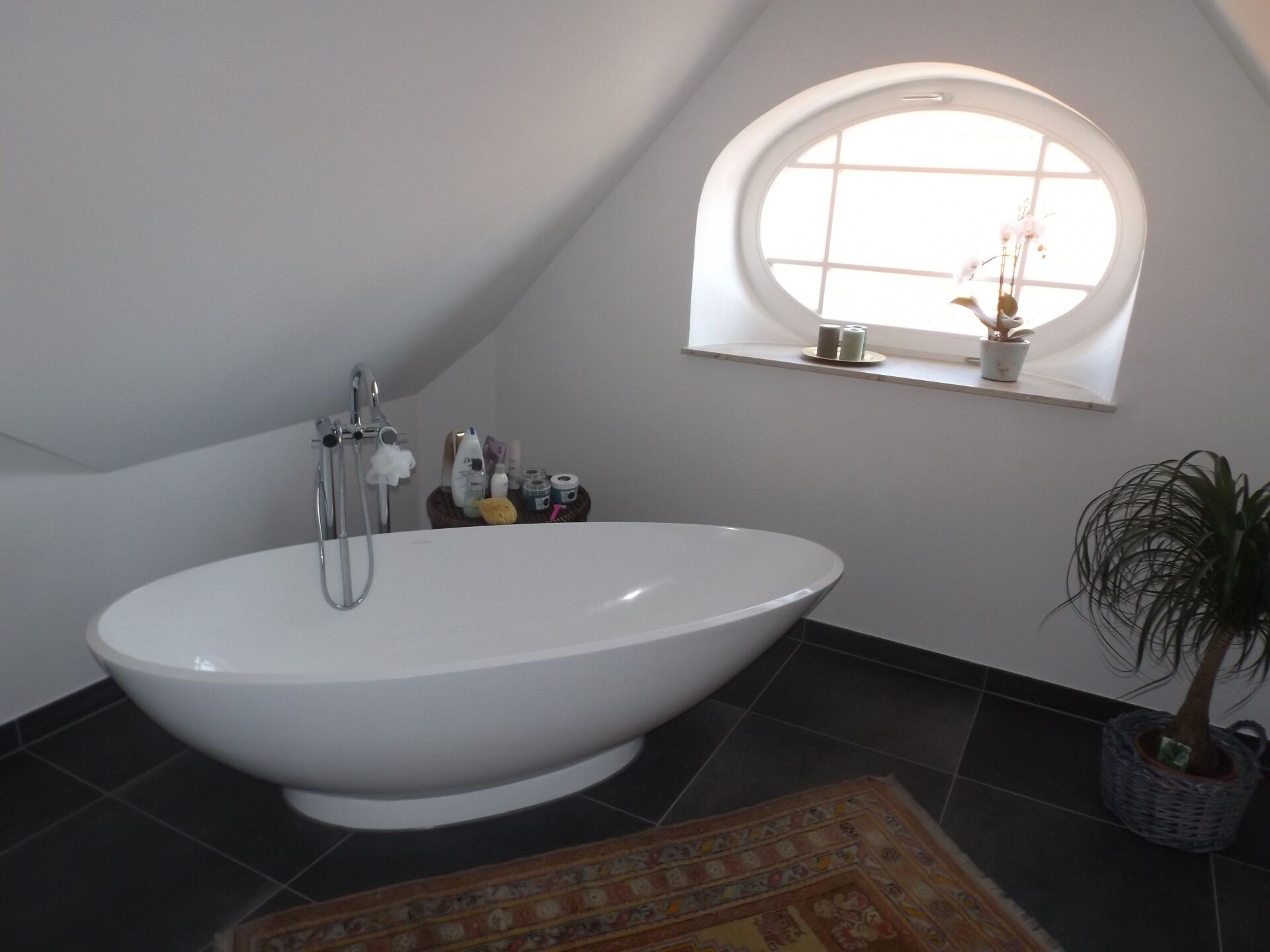 Ovale Badewanne in einem weiß getünchten Badezimmer mit Bogenfenster, dunklem Fliesenboden und kleinem Teppich.