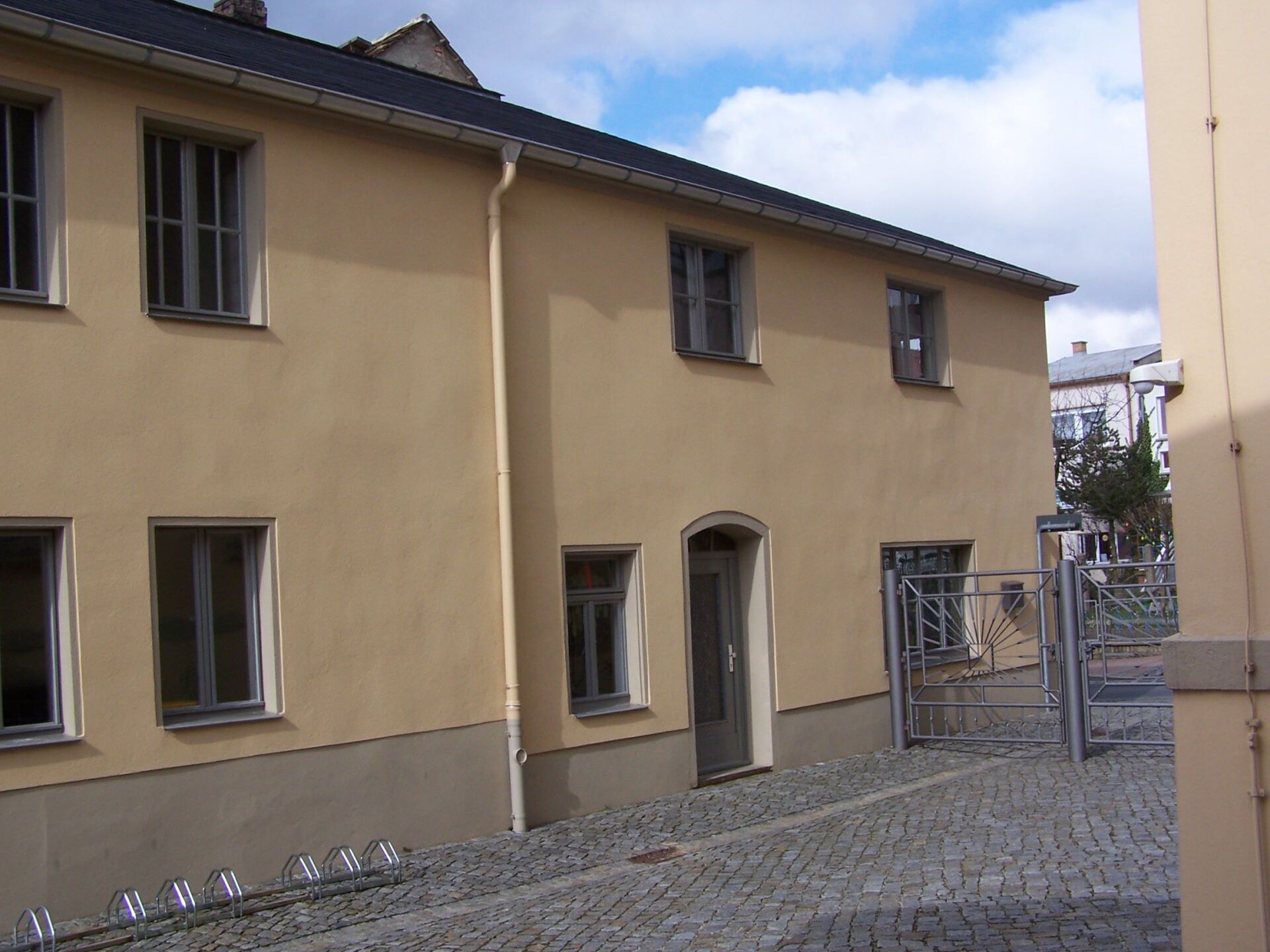 Beiges Gebäude mit mehreren Fenstern und einer Tür, Kopfsteinpflasterstraße, blauer Himmel mit Wolken.