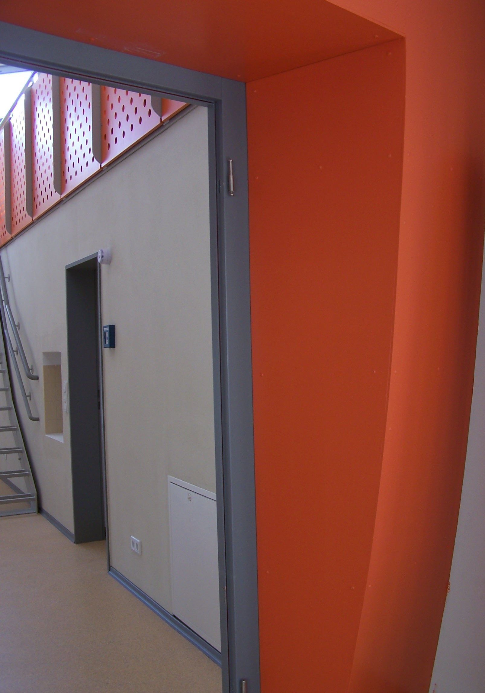 Eine orangefarbene Wand neben einem grauen Türrahmen führt zu einem Flur mit Treppenhaus und Aufzug.