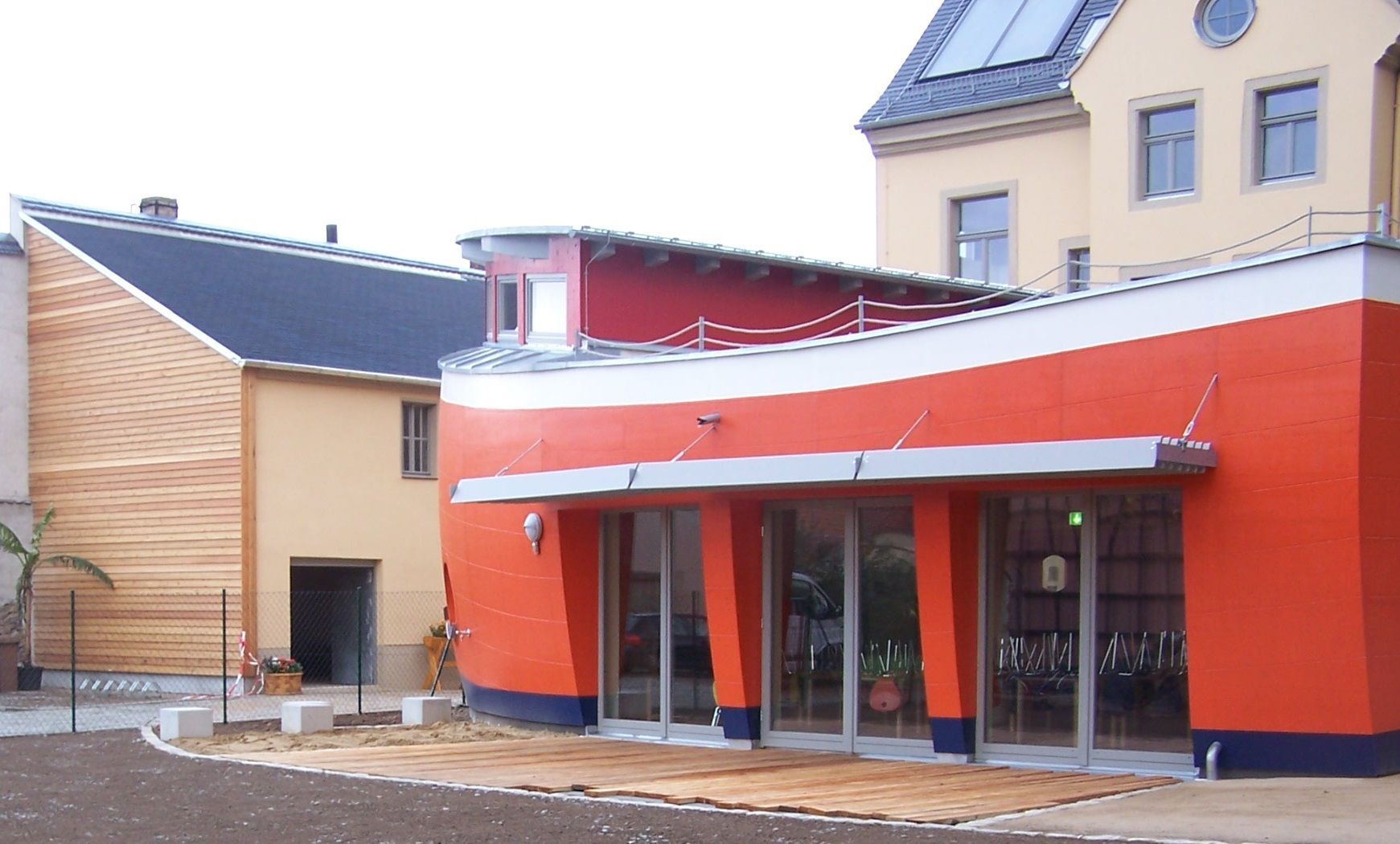 Ein orange-blaues, geschwungenes Gebäude mit Glastüren steht neben einem Gebäude mit Holzverkleidung.