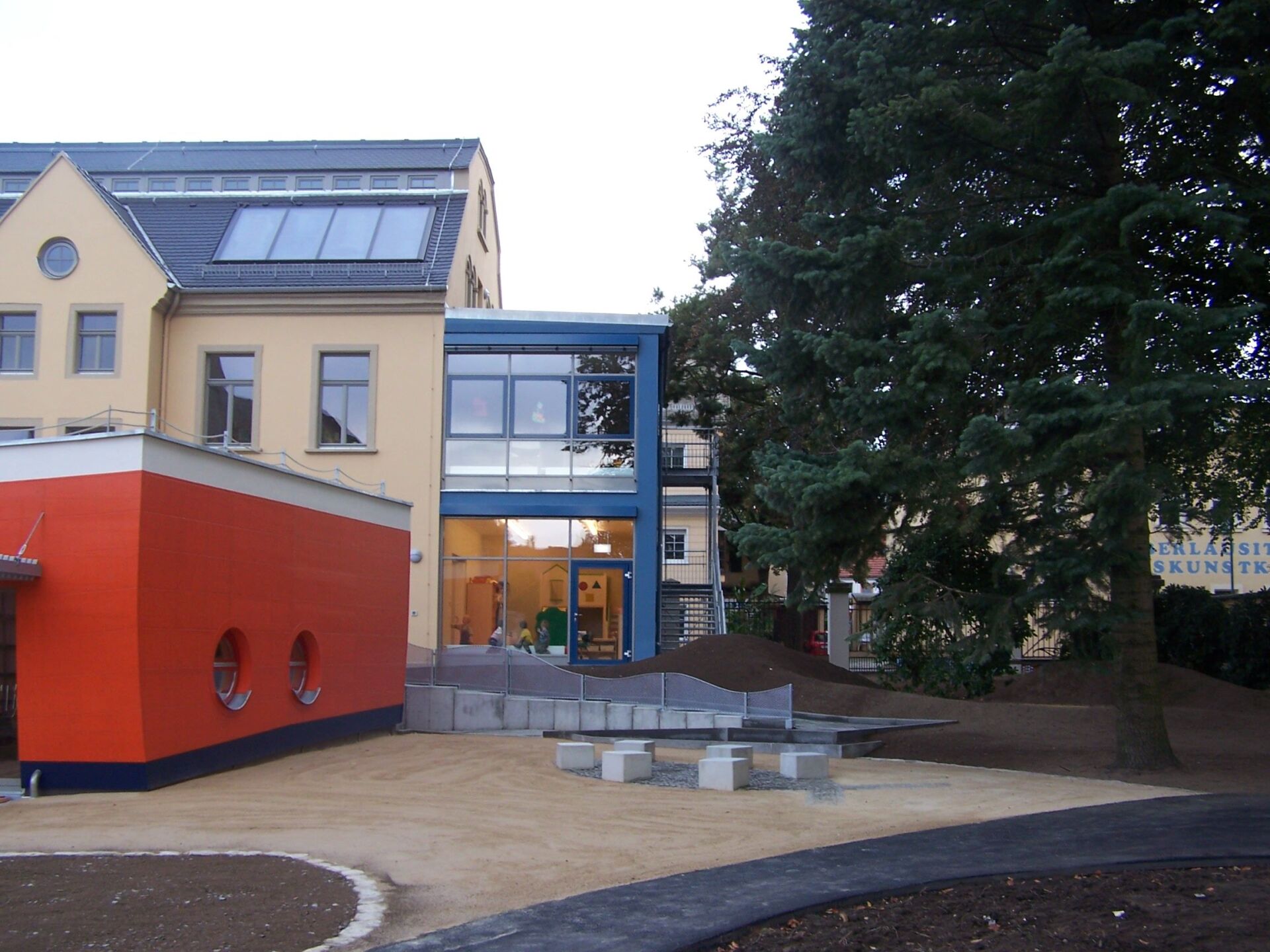 Farbenfrohes Gebäude mit roten und gelben Abschnitten, Glaseingang, Bäumen und kleinem Spielplatz.