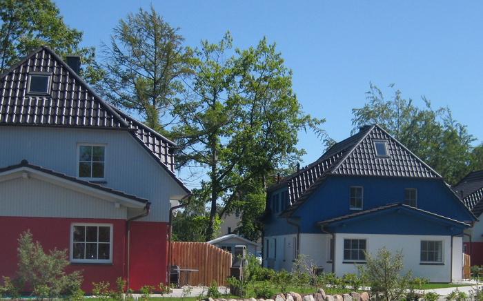 Zweistöckige Häuser mit roten, blauen und weißen Fassaden, schwarzen Ziegeldächern und umgeben von Bäumen unter blauem Himmel.