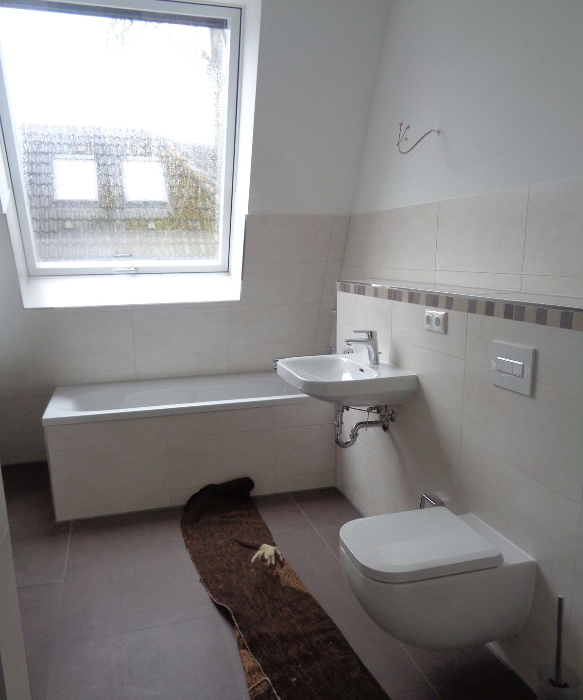Badezimmer mit weißen Armaturen: Badewanne, Waschbecken und Toilette, neben einem Fenster. Brauner Teppich auf dem Boden, neutrale Wände.