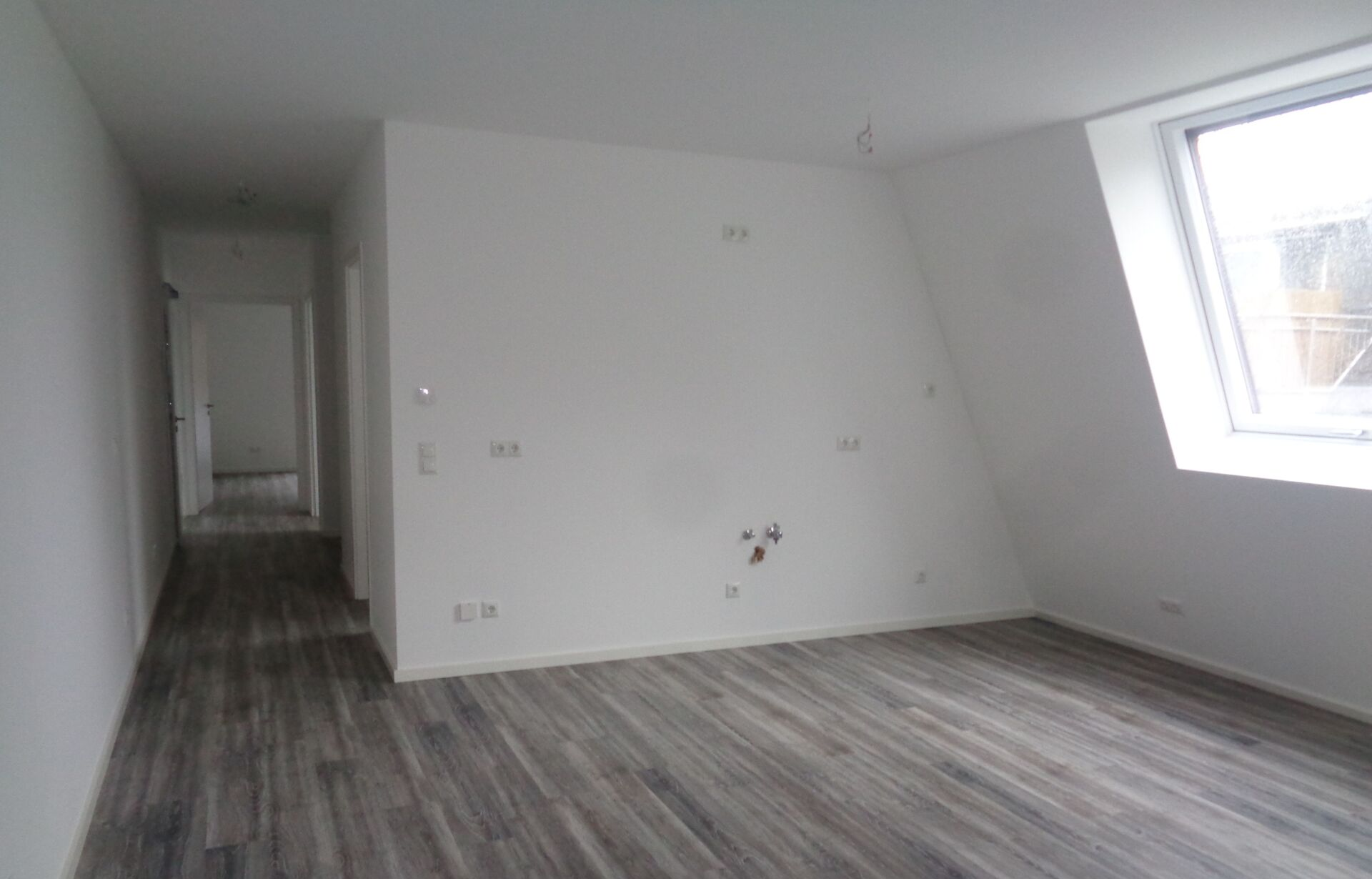 Leerer Raum mit grauem Holzboden, weißen Wänden und einem Fenster auf der rechten Seite.