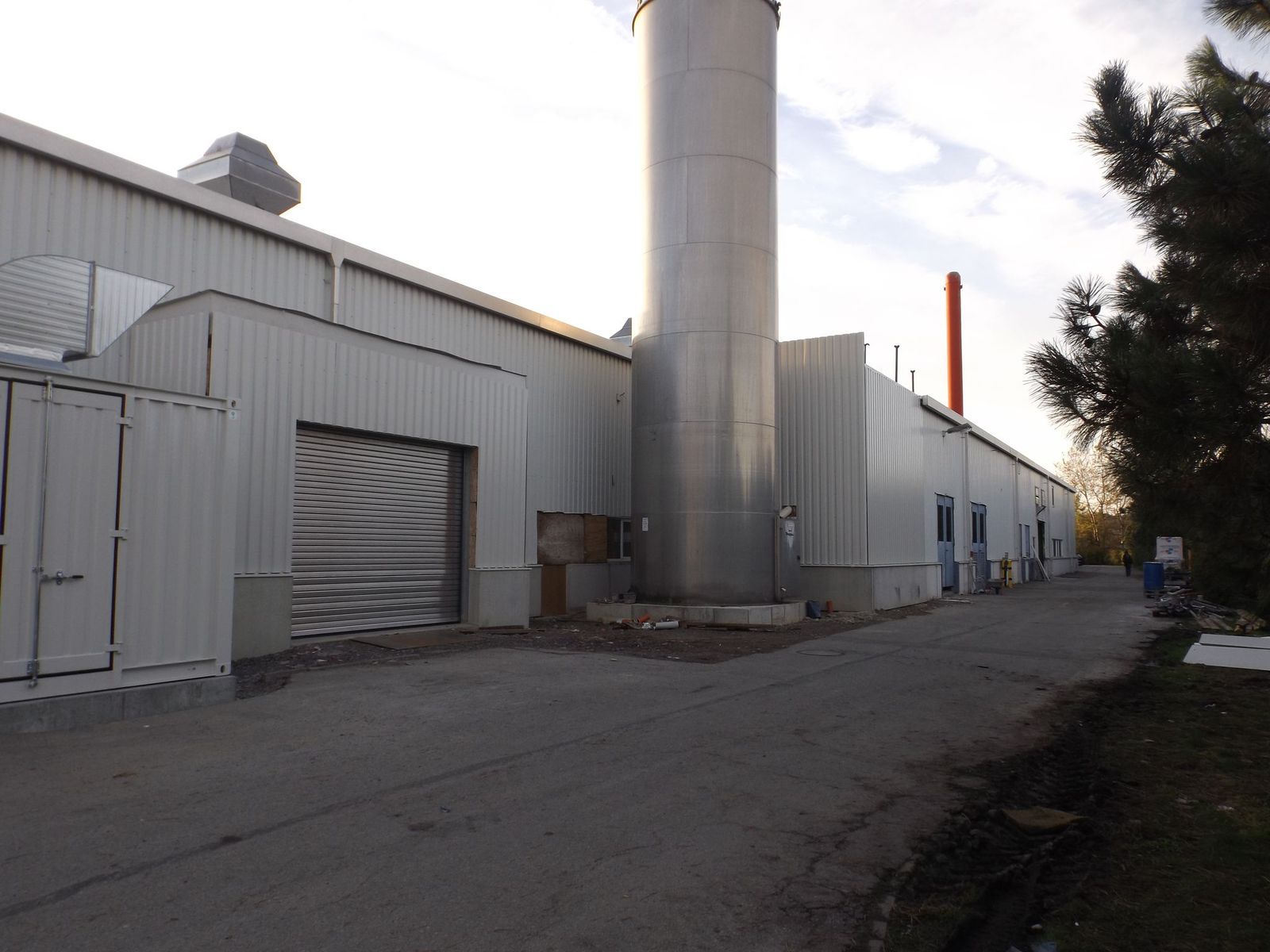 Industriegebäude mit großem Schornstein, Rolltor und Metallverkleidung. Gepflasterter Bereich davor.