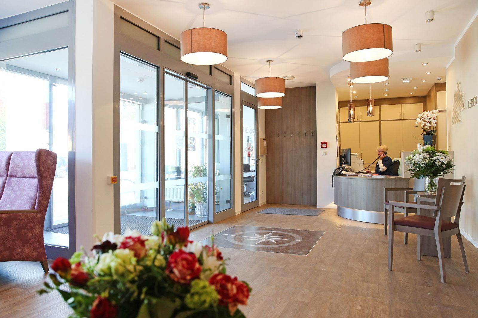 Lobby mit automatischen Türen, Rezeption, Topfblumen und Pendelleuchten. Eine Person sitzt am Empfangstresen.