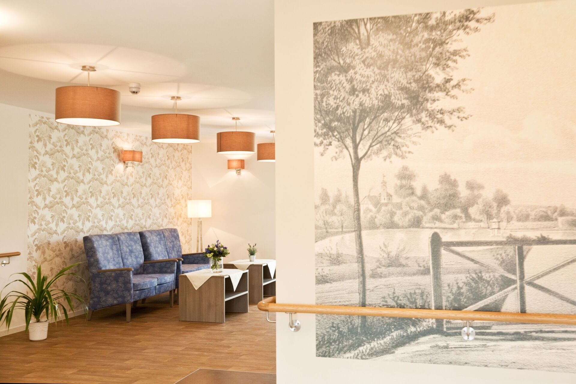 Lounge-Bereich mit blauem Sofa, Tischen, dekorativer Tapete und großem Wandgemälde einer ländlichen Landschaft. Braune Pendelleuchten.