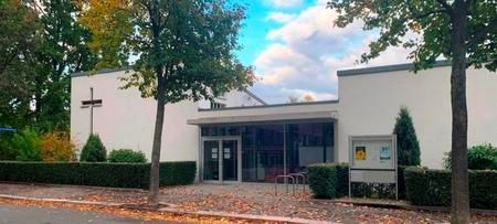 Ein modernes Kirchengebäude mit Kreuz, Bäumen und einer Informationstafel vor bewölktem Himmel.