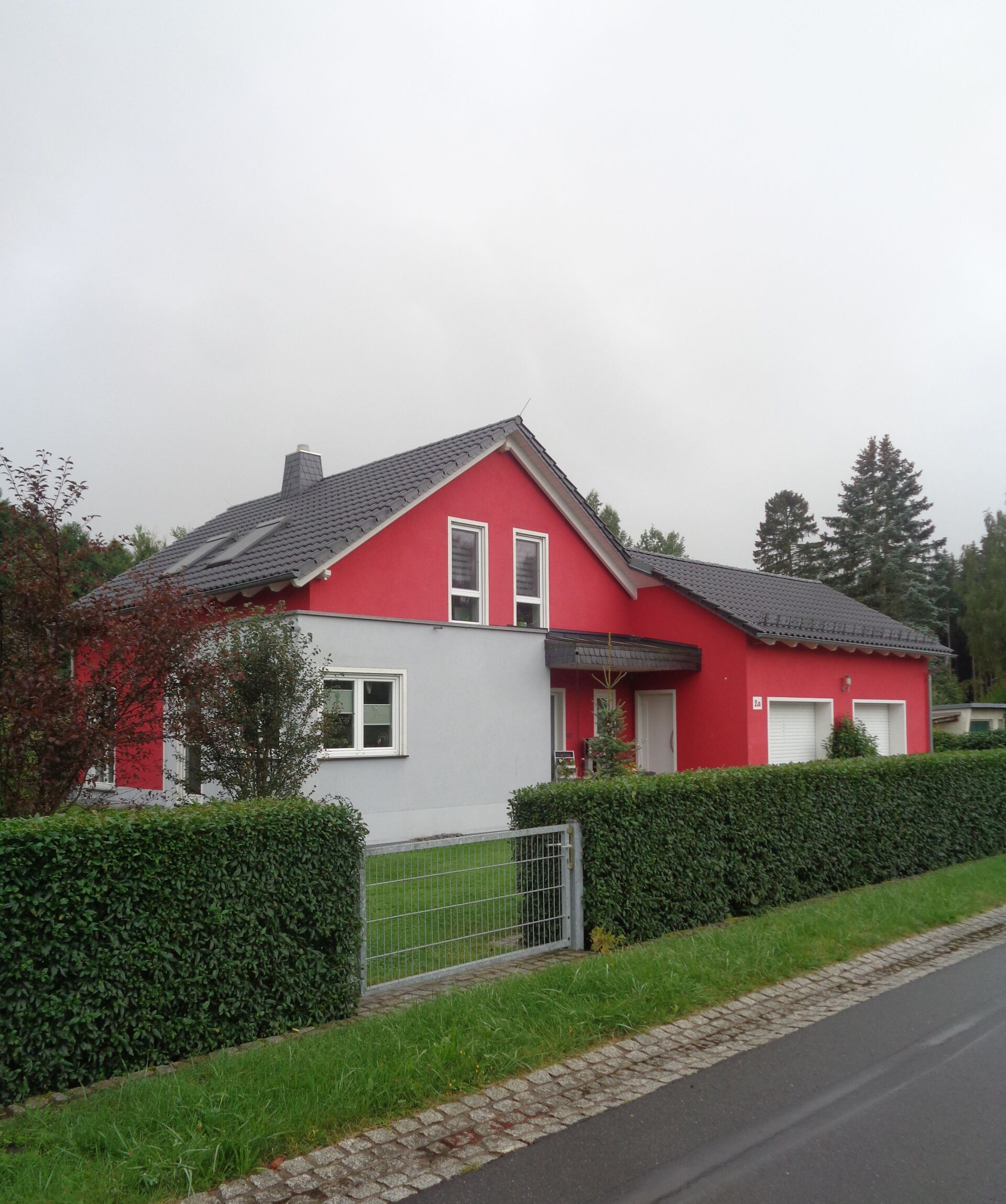 Rot-graues Haus mit schwarzem Dach, weißen Zierelementen und einer Hecke davor.