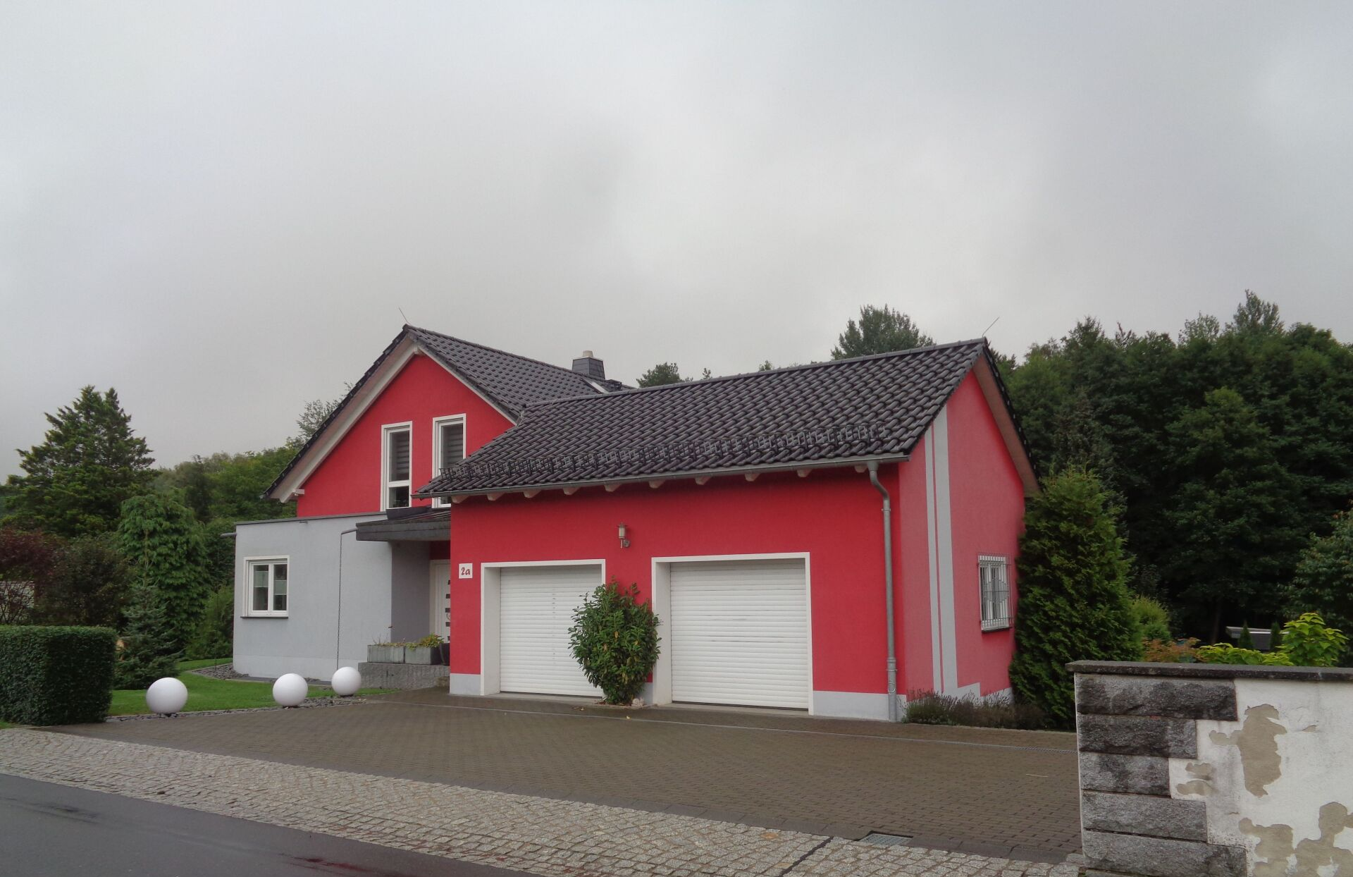 Rotes Haus mit Garage, grauen Zierelementen und weißen Garagentoren unter bedecktem Himmel.