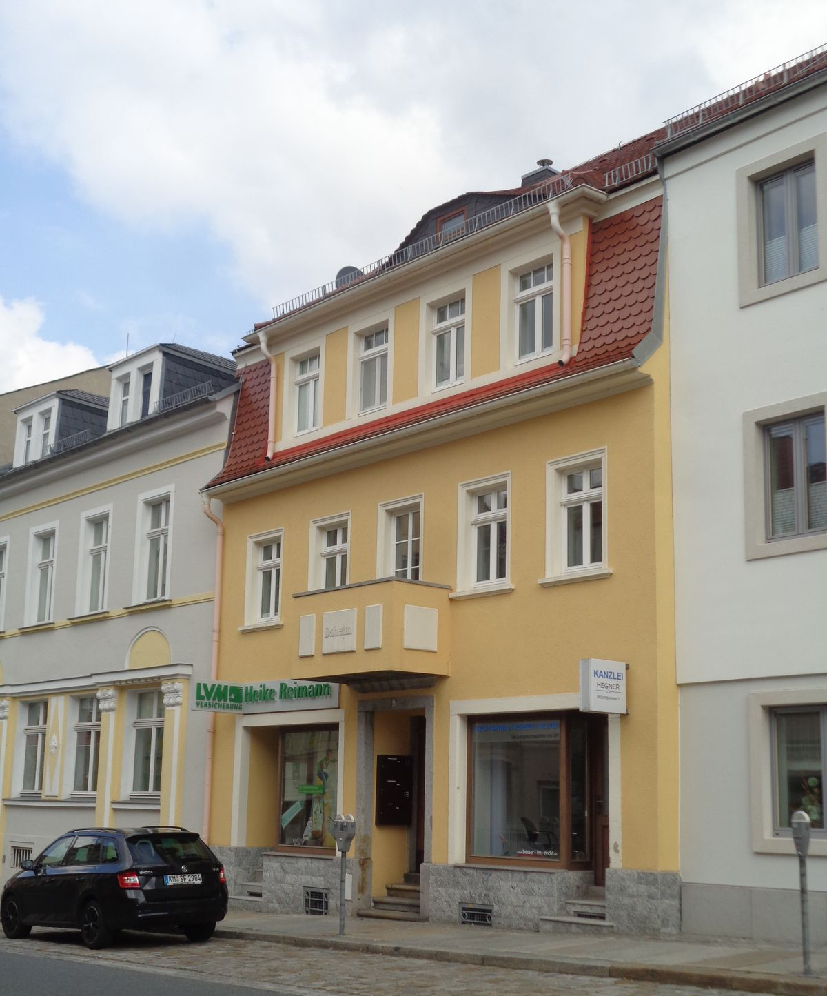Gelbes Gebäude mit Ladenfronten und mehreren weiß gerahmten Fenstern, rotem Ziegeldach, geparktem Auto, Backsteinstraße.