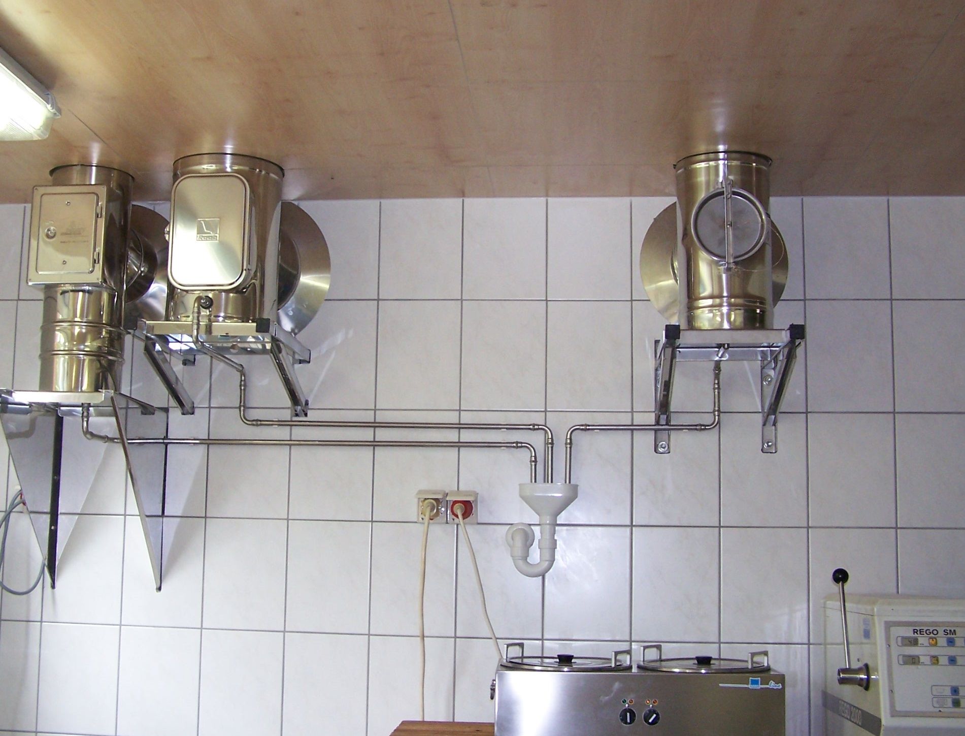 Verarbeitungsanlagen aus Edelstahl, montiert an einer gefliesten Wand, mit Verbindungsrohren und einem zentralen Abfluss.